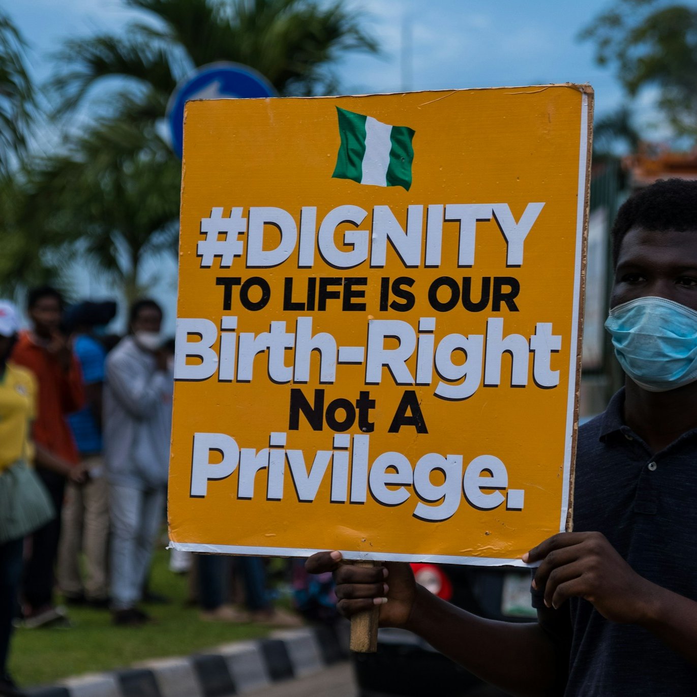 Sur l'image, une manifestation a lieu dans la rue. Un homme se tient au premier plan, portant un masque. Il tient une grande pancarte colorée qui affiche le message suivant : "#DIGNITY TO LIFE IS OUR Birth-Right Not A Privilege" en lettres grandes et noires sur un fond orange. En arrière-plan, on aperçoit un groupe de personnes, certaines portant également des masques, rassemblées autour de lui. L'environnement est urbain, avec des arbres et des véhicules visibles. L'atmosphère est engagée, témoignant d'une lutte pour les droits et la dignité humaine.