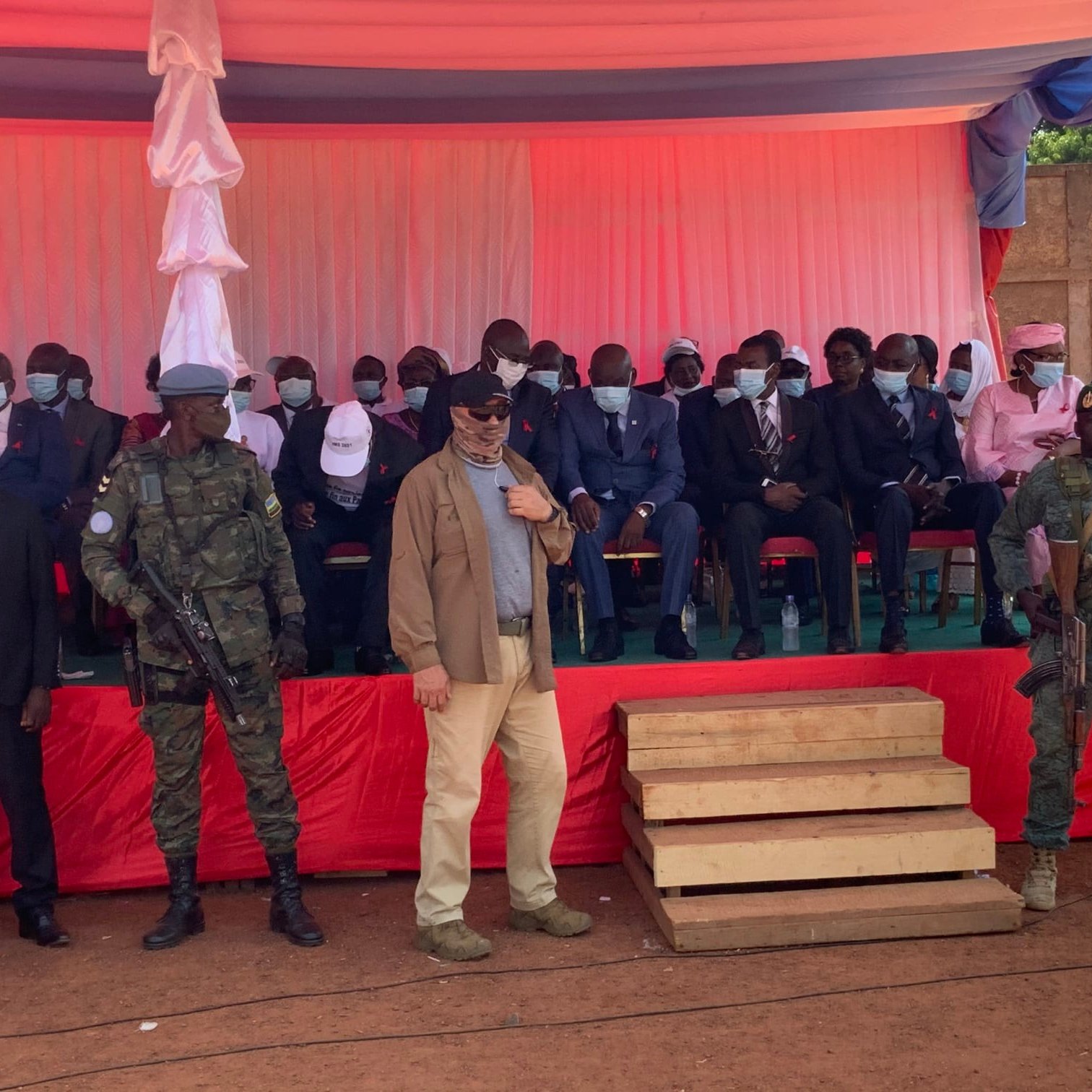 L'image montre une scène d'événement en plein air, probablement une cérémonie officielle. Au premier plan, deux soldats en uniforme, l'un avec un casque et l'autre portant une veste militaire, se tiennent debout à côté de deux marches en bois. Derrière eux, un groupe de personnes est assis sur des chaises, en grande partie habillées de manière formelle, certaines portant des masques. Les hommes et les femmes sont alignés en rang, sur un fond de tissu rouge et blanc. L'ambiance semble solennelle, avec des drapeaux et des décorations typiques des événements institutionnels. Le décor est éclairé par une lumière naturelle, et l'atmosphère pourrait évoquer une cérémonie de discours ou une célébration publique.