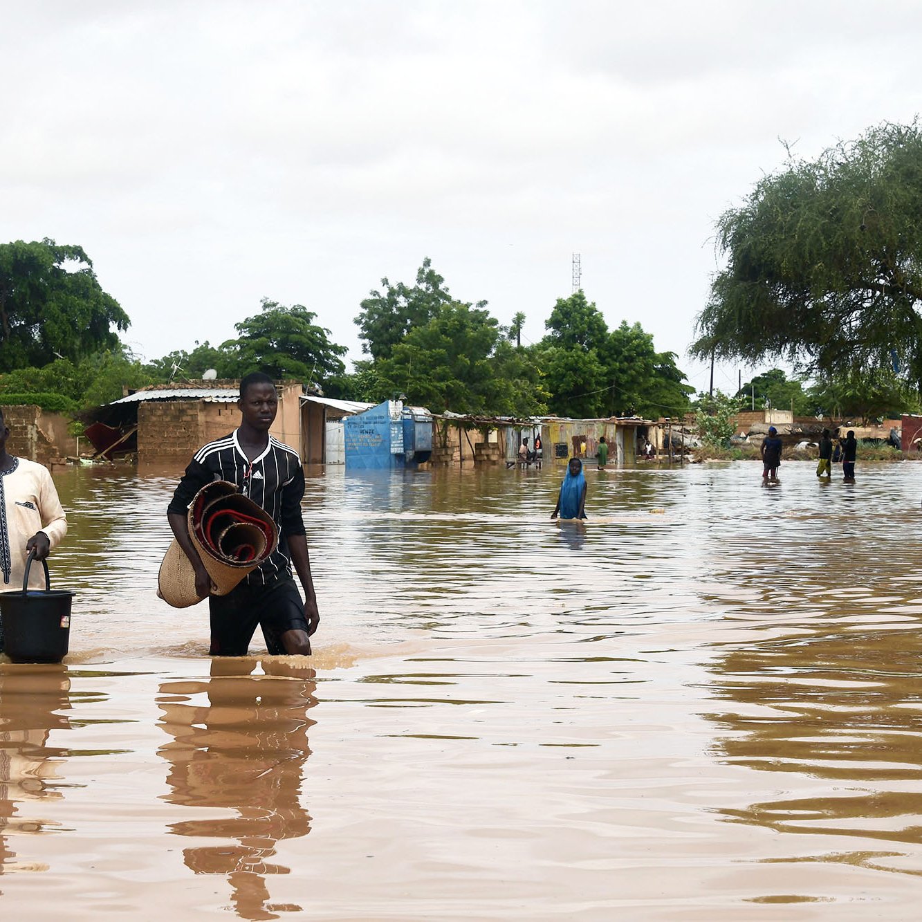 The image depicts a flooded area, with individuals wading through water that has inundated the surroundings. The water reaches knee-deep, affecting homes and structures in the background, which appear to be partially submerged. People in the scene are carrying various items, suggesting they're either evacuating or salvaging belongings. Trees and greenery can be seen around the area, indicating that it is a rural or semi-urban environment. The sky is overcast, contributing to the somber atmosphere of the flooding situation.
