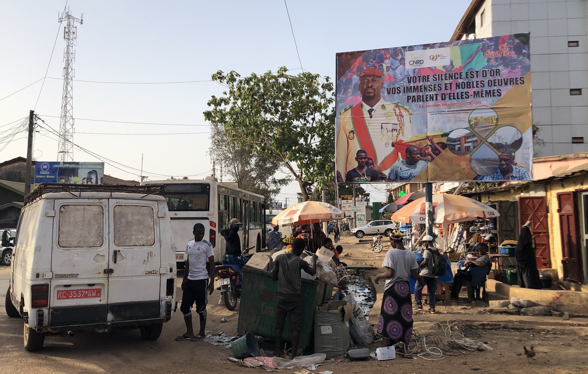 L'image représente une scène de rue animée dans une ville. Au premier plan, on voit une camionnette blanche à l'arrière, avec des détails usés et des portes ouvertes. À gauche, un bus de transport en commun est stationné. Plusieurs personnes se déplacent sur la chaussée, certaines vendant des marchandises sous des parasols colorés. Il y a également des bacs de récupération et divers déchets sur le sol, indiquant une ambiance de marché. En arrière-plan, un grand panneau publicitaire présente des visages de personnes et un message en français, affichant un slogan qui semble inciter à l'action ou au changement. Les arbres et les fils électriques ajoutent à l'atmosphère urbaine du lieu.