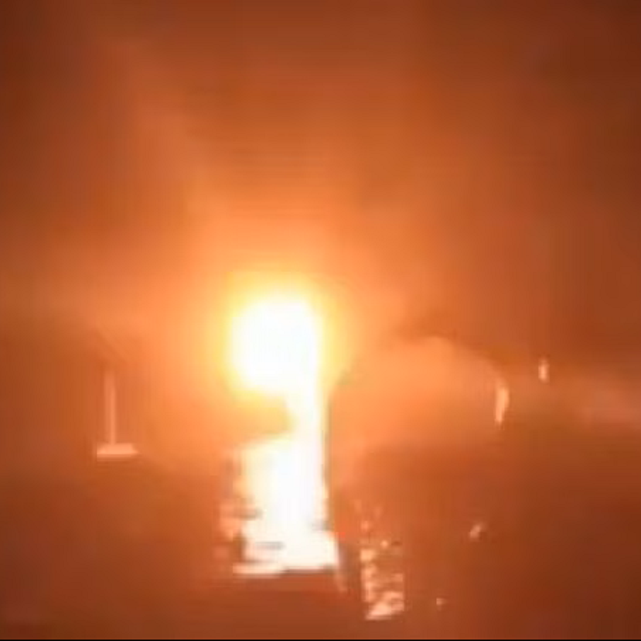 L'image présente une scène sombre avec une source de lumière éclatante au centre, semblant provenir d'un feu ou d'un soleil couchant. Les contours des objets et des formes sont flous, créant une ambiance mystérieuse. L'environnement pourrait évoquer un paysage naturel ou urbain, sans détails précis visibles. On ressent une intensité et une chaleur émises par cette lumière, contrastant avec l'obscurité environnante.