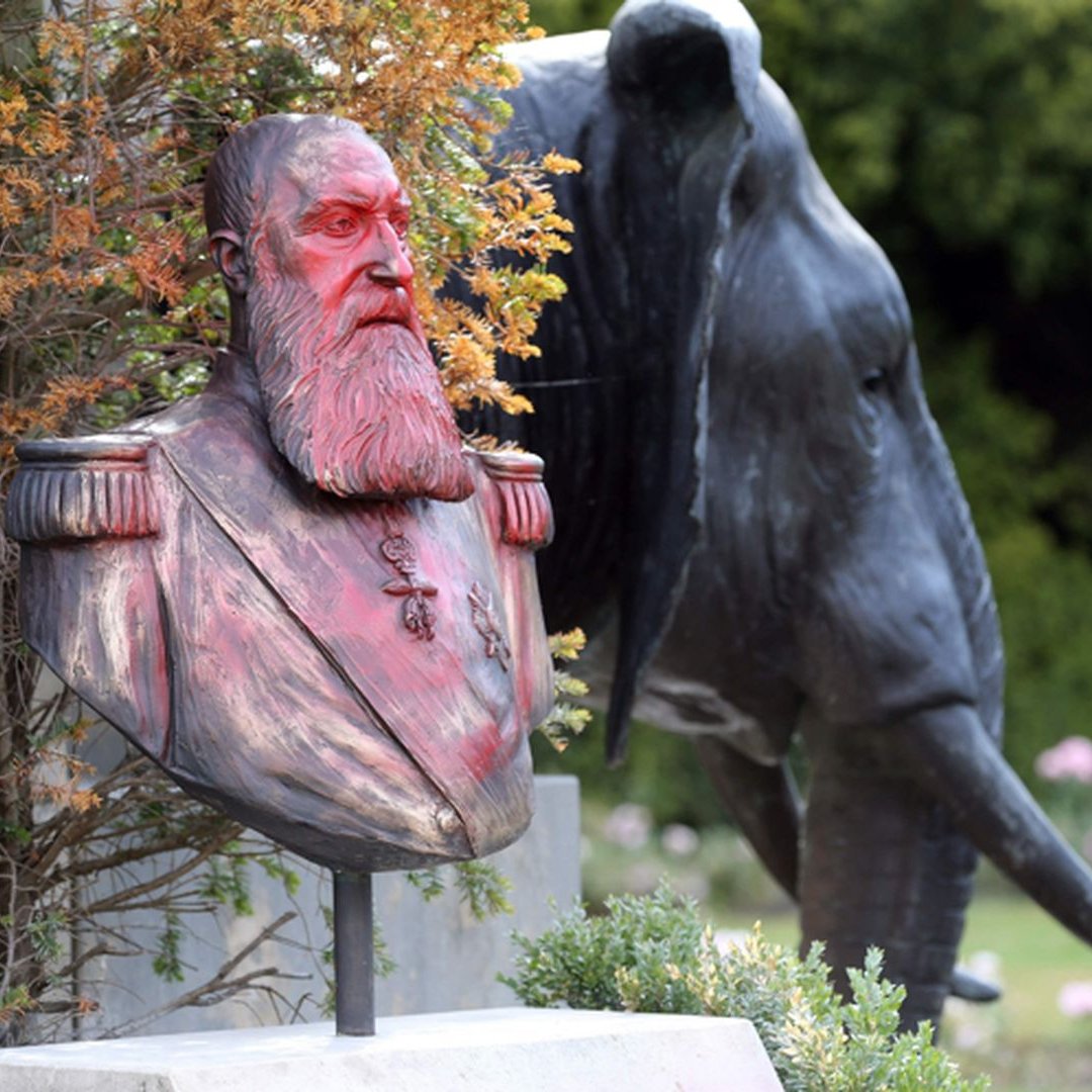 L'image présente un buste en bronze d'un homme barbu, aux cheveux longs et portant des uniformes militaires, posé sur un socle. Ce buste est entouré de végétation, ce qui lui donne un aspect naturel. En arrière-plan, on aperçoit la silhouette d'un éléphant en métal, affichant des détails réalistes comme ses grandes oreilles et sa trompe. Le contraste entre le buste humain et l'éléphant, ainsi que la verdure environnante, crée une ambiance paisible et mémorable dans un jardin.