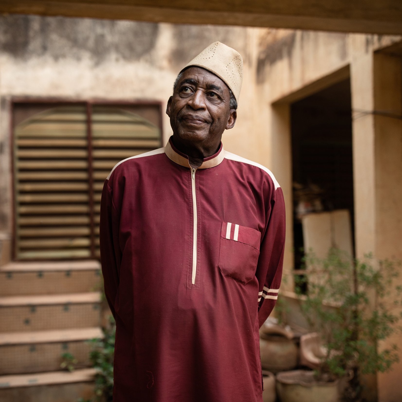 L'image montre un homme âgé portant une tenue traditionnelle d'une couleur rouge foncé. Sa chemise longue est agrémentée de détails blancs. Il se tient debout, les bras le long du corps, et regarde légèrement vers le haut. L'arrière-plan présente un bâtiment avec des murs usés et une porte en bois, ainsi que quelques plantes verdoyantes qui ajoutent une touche de nature à la scène. L'homme dégage une expression calme et réfléchie, créant une atmosphère de sagesse et de sérénité.