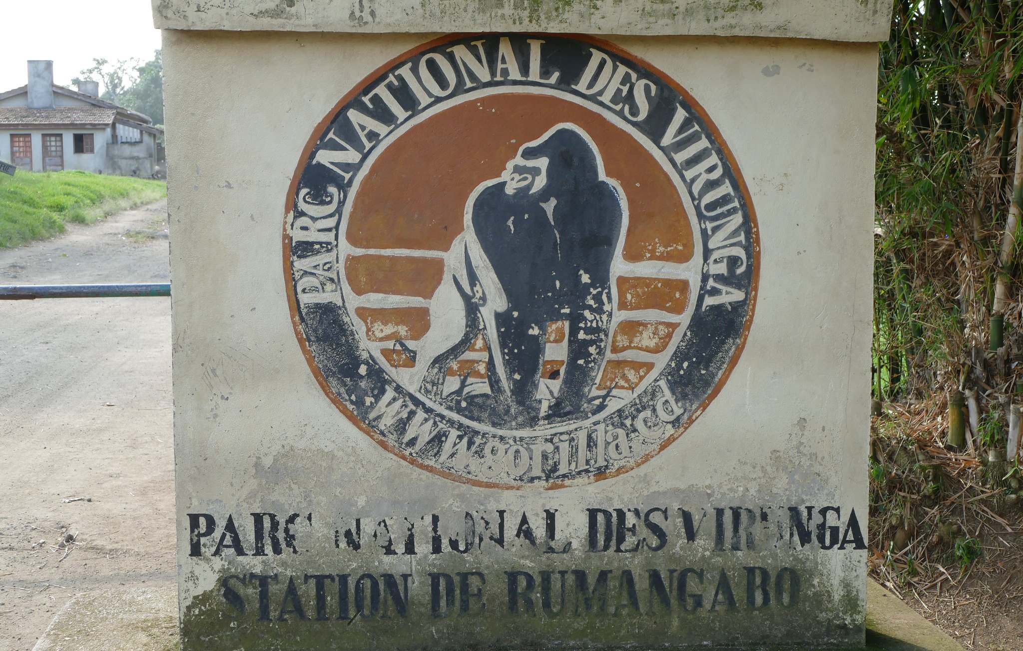 L'image montre un panneau en béton qui indique l'entrée du Parc National des Virunga. Le panneau est décoré d'un logo représentant un gorille, qui est l'un des emblèmes du parc. Les mots "PARC NATIONAL DES VIRUNGA" et "STATION DE RUMANGABO" sont inscrits en lettres claires sur la surface du panneau. En arrière-plan, on peut apercevoir des bâtiments et une route qui semble mener vers la nature environnante. L'ensemble de la scène évoque une atmosphère d'aventure et de préservation de la faune.