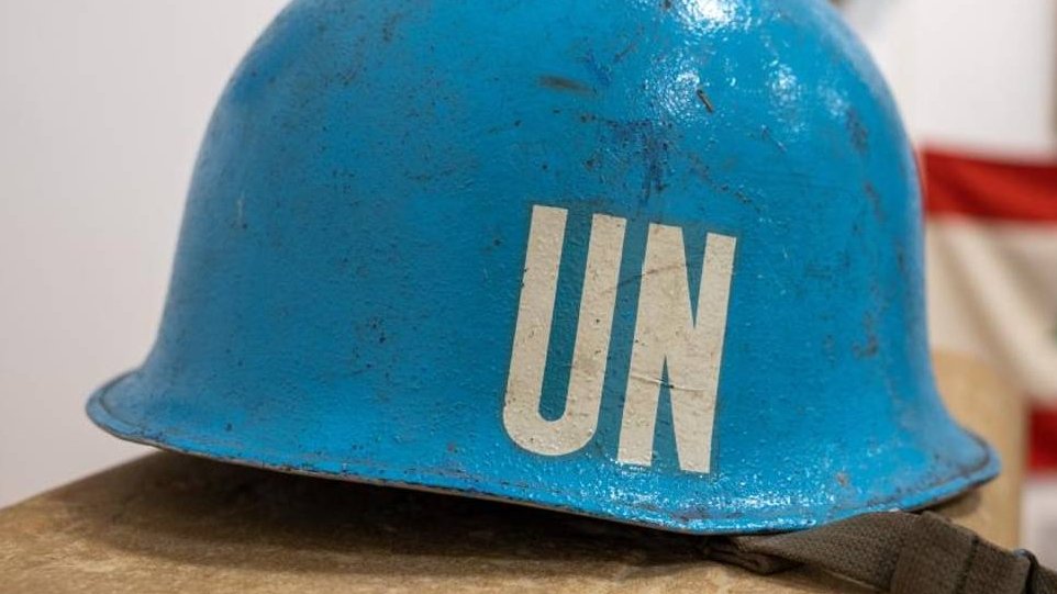 L'image montre un casque de couleur bleu vif, typique des casques de l'ONU. La surface du casque est légèrement abîmée, indiquant qu'il a probablement été utilisé sur le terrain. Sur le devant, des lettres blanches "UN" sont bien visibles, représentant l'Organisation des Nations Unies. Le casque est posé sur une surface plane, avec un fond flou qui évoque un environnement de musée ou d'exposition, où des objets historiques peuvent être présentés. Ce casque symbolise la paix et les missions de maintien de la paix menées par l'ONU dans diverses régions du monde.