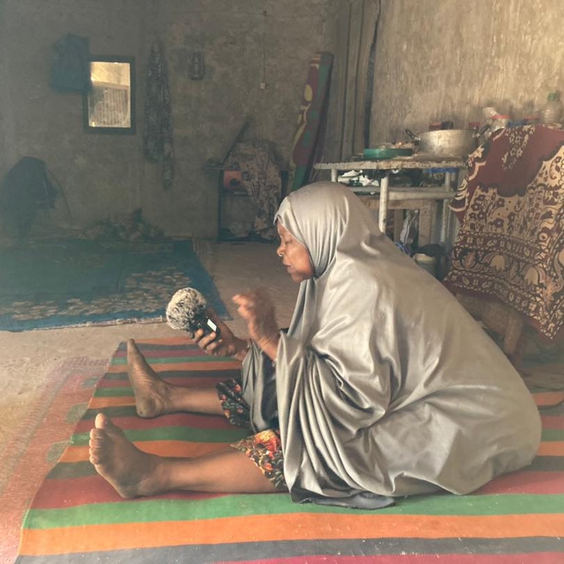 L'image représente une femme assise sur un tapis coloré dans un intérieur simple et rustique. Elle porte un vêtement ample et gris, qui couvre son corps. Sa posture est attentive, et elle tient un petit objet dans ses mains, probablement un instrument de musique ou une pièce d'artisanat. L'environnement autour d'elle est dépouillé, avec des murs en béton et quelques éléments de décoration, comme des tapis et des coussins. On peut sentir une atmosphère calme et intime, presque spirituelle. Des rayons de lumière pénètrent à travers une fenêtre, illuminant doucement la scène.