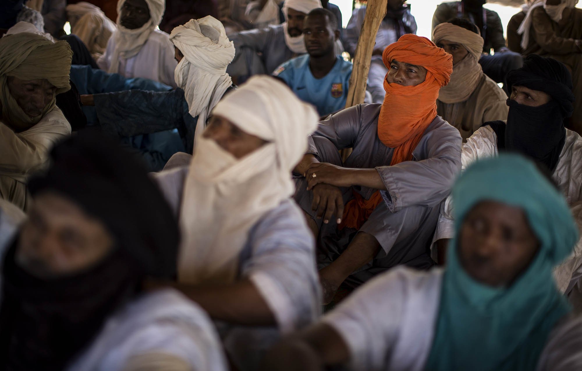 L'image montre un groupe de personnes assises dans un espace clos. Les individus portent des vêtements traditionnels, souvent en tissu léger, et sont partiellement masqués par des turbans ou d'autres pièces de tissu. On peut percevoir des couleurs variées : certains portent des turbans orange, d'autres bleus ou verts. Les visages des personnes sont en grande partie cachés, créant une atmosphère de mystère. L'environnement semble calme, et les positions des corps indiquent une attention collective. La lumière est douce, suggérant un éclairage naturel venant d'une source extérieure. On peut également sentir une ambiance de rassemblement, possiblement pour un échange culturel ou une discussion importante.