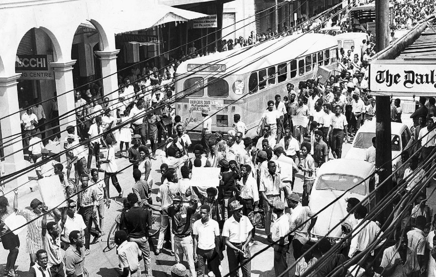 L'image que vous avez devant vous représente une scène urbaine animée dans les années 1960. On peut voir une foule dense de personnes marchant dans une rue. Les gens portent des vêtements typiques de l'époque, et il semble qu'ils se déplacent avec un but précis. À gauche, plusieurs commerces sont visibles, avec des enseignes en lettres visibles. Un ancien bus à l'allure caractéristique est garé, ajoutant une touche nostalgique à la scène. On aperçoit également quelques voitures stationnées et d'autres qui circulent dans la rue. Les fils électriques sont visibles en haut de l'image, accentuant l'impression d'une ville vivante et dynamique. L'atmosphère générale évoque une période de mouvement socioculturel important, avec des échanges et une vitalité palpable dans l'air.