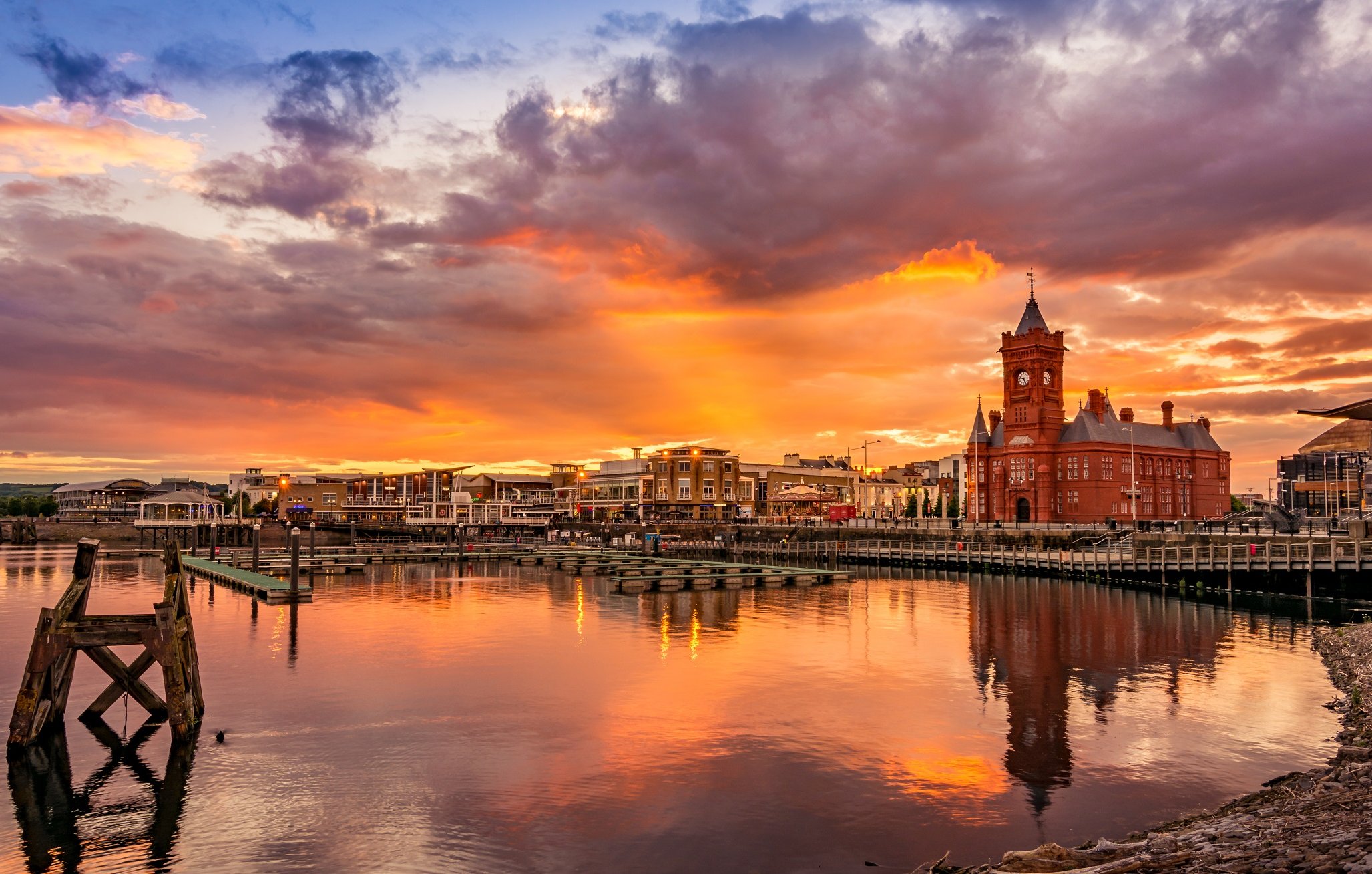 Cette image représente un paysage de bord de mer au coucher du soleil. À l'horizon, un ciel splendide est illuminé par des teintes enflammées de rouge, orange et violet. Le soleil déclinant éclaire les nuages, créant une atmosphère magique. Au premier plan, l'eau calme d'une marina reflète ces couleurs vives, tandis que plusieurs quais et petits bateaux ajoutent à la scène paisible. Sur la rive, un bâtiment architectural distinctif de couleur rouge, avec une tour pointue, attire l'attention, tandis que des arbres et des éléments naturels encadrent le paysage, offrant une ambiance sereine et accueillante.