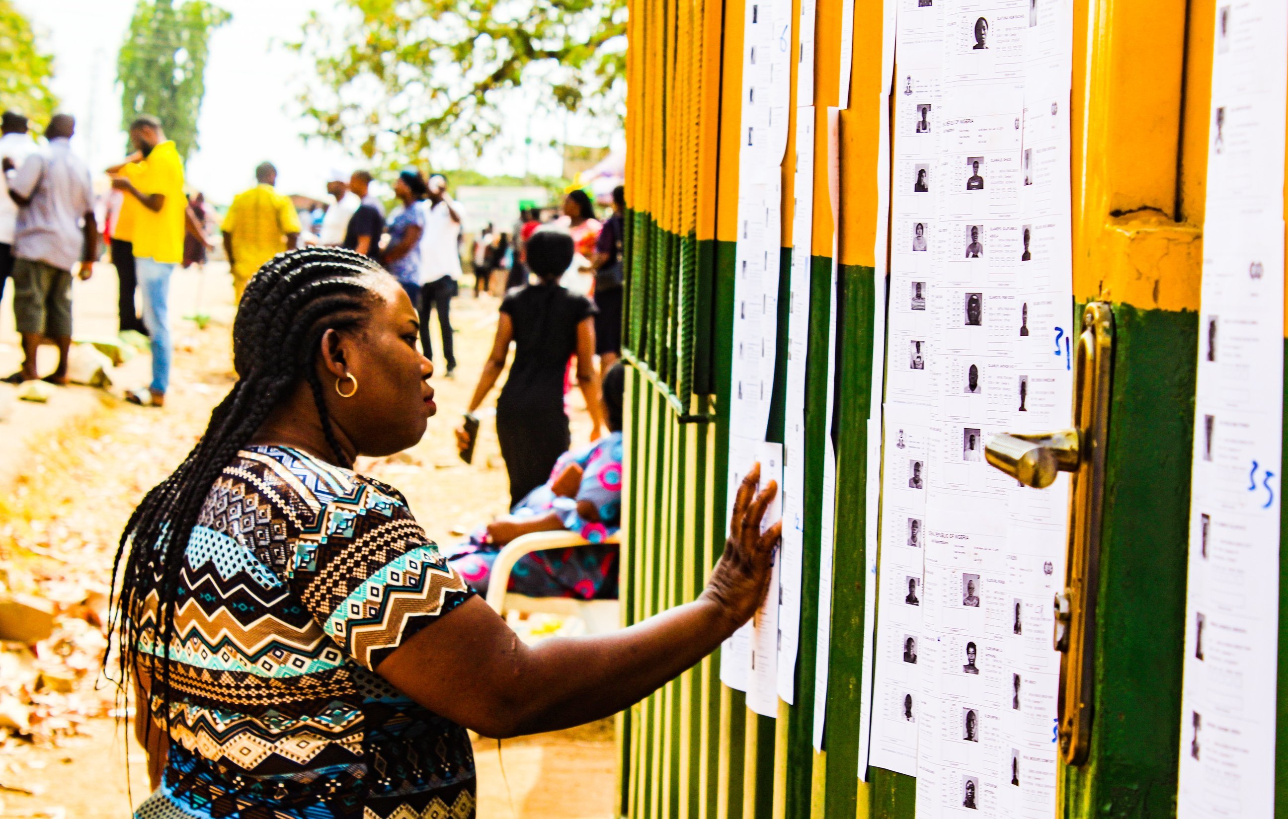 Dans cette image, on voit une scène animée en extérieur. Une femme, avec des tresses et portant un haut aux motifs colorés, se tient près d'un mur recouvert de plusieurs feuilles de papier. Elle passe sa main sur l'une de ces feuilles, qui semble contenir des informations, probablement des listes ou des résultats. En arrière-plan, de nombreuses autres personnes se déplacent, créant une atmosphère de rassemblement. Les couleurs vives du mur et la lumière du jour ajoutent une ambiance chaleureuse à la scène. Au sol, on peut apercevoir des feuilles mortes, ce qui suggère une saison automnale.