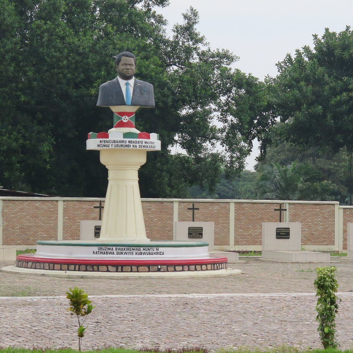 Cette image montre une statue en l'honneur d'une personnalité importante, probablement un leader ou un héros national. La statue est placée sur un socle élevé et représente un homme, vêtu d'un costume. En arrière-plan, on peut voir un mur avec plusieurs croix, qui semblent être des stèles commémoratives. Le site est entouré de verdure, avec des arbres touffus qui apportent une atmosphère paisible. Le sol est en pavé, et des plantes décoratives sont présentes, ajoutant une touche de nature à cet espace de mémoire.