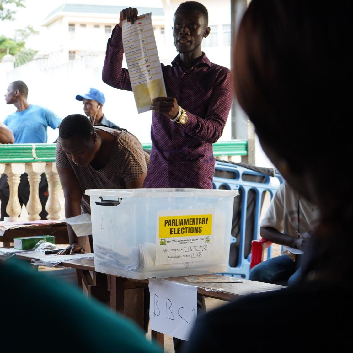 L'image montre une scène d'élections parlementaires. Au centre, un homme se tient debout, levant une feuille de papier, probablement un bulletin de vote, pour montrer aux personnes présentes. Il s'habille d'une chemise de couleur sombre et semble expliquer quelque chose de manière engageante. En arrière-plan, plusieurs personnes sont rassemblées, certaines observant attentivement tandis que d'autres attendent, adossées à une balustrade. On devine une atmosphère de concentration et d'anticipation, typique d'un moment électoral. Sur une table, une boîte transparente étiquetée "ELECTIONS PARLEMENTAIRES" est visible, contenant probablement des bulletins de vote. 