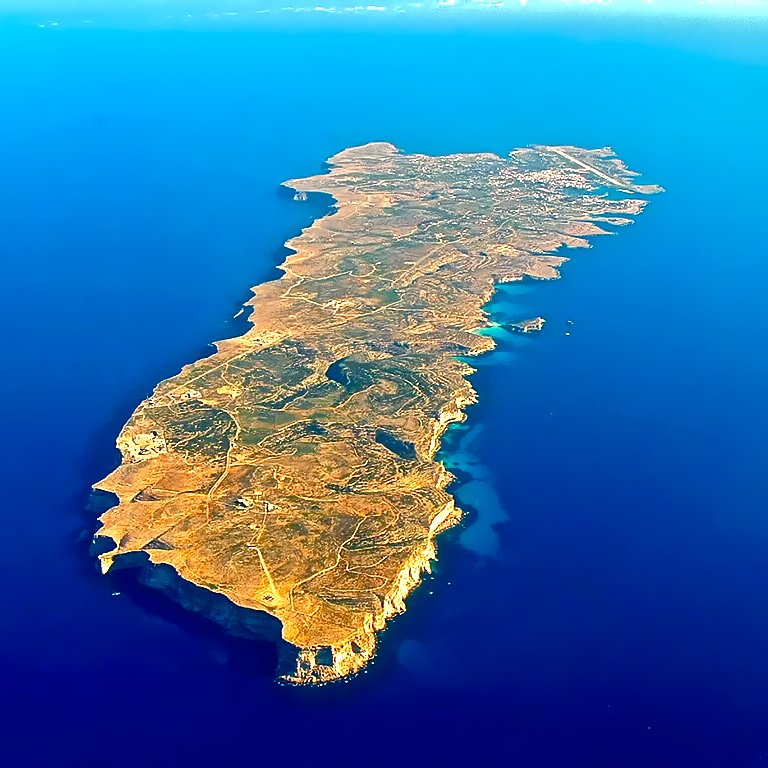 L'image représente une île vue du ciel, entourée par une vaste étendue d'eau bleue. L'île a une forme allongée et irrégulière, avec des côtes découpées et quelques zones rocheuses. Sa surface est en grande partie couverte de végétation, avec des teintes de jaune, de vert et de brun qui évoquent un terrain sec et accidenté. On peut également observer quelques petites baies et criques où l'eau semble se jeter sur le rivage. L'ensemble évoque une atmosphère de tranquillité et de beauté naturelle, typique d'une destination insulaire.