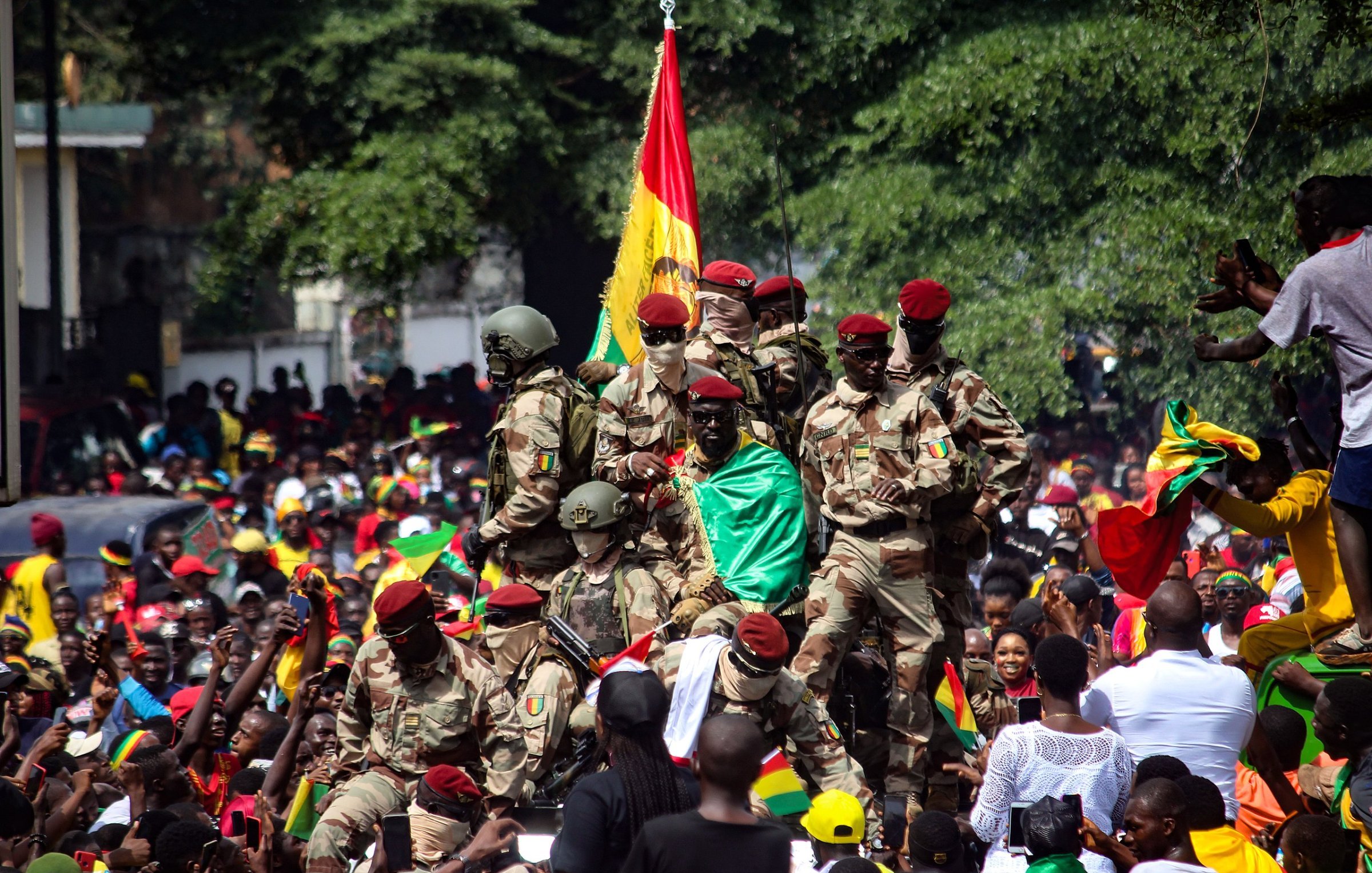 L'image montre une scène de célébration animée. Au premier plan, un groupe de soldats en uniforme militaire se tient debout, certains brandissant un drapeau aux couleurs vives de la Guinée, rouge, jaune et vert. Ils semblent être au centre d'une foule massive, qui est remplie de personnes qui portent également des vêtements aux couleurs du drapeau. L'ambiance est festive et euphorique, avec des visages souriants et levés vers les soldats. En arrière-plan, on aperçoit des arbres et des bâtiments, créant un cadre urbain. L'énergie collective de la foule et la présence militaire suggèrent un événement significatif, probablement une célébration ou une manifestation patriotique.