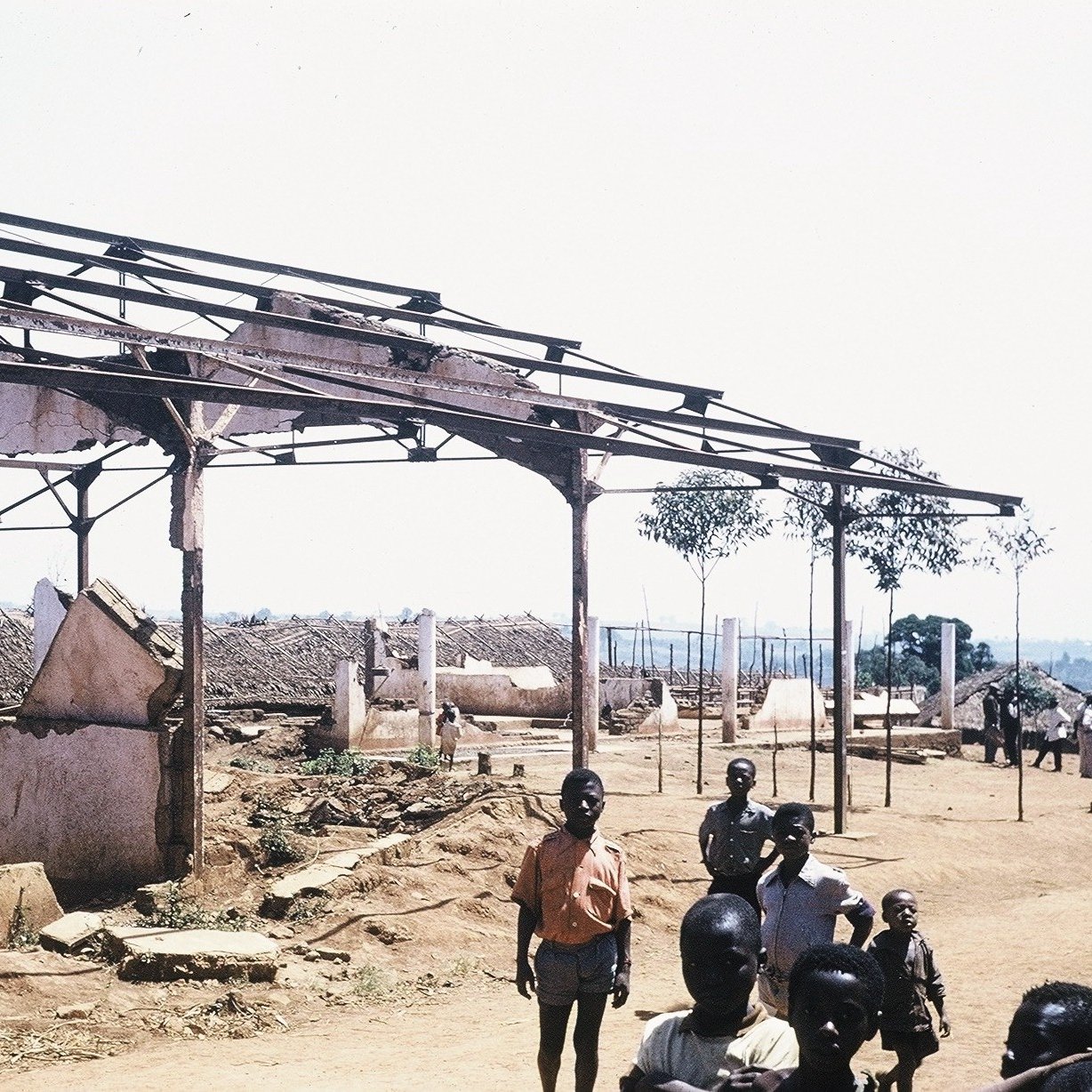 L'image montre un paysage de dévastation avec des structures en ruine. À l'avant-plan, plusieurs personnes, dont des enfants et des adultes, se tiennent debout sur un sol poussiéreux. En arrière-plan, on voit des bâtiments partiellement effondrés, avec des toits manquants et des murs endommagés. Des arbres isolés se dressent ici et là, ajoutant une touche de verdure à ce cadre désolé. Le ciel est dégagé et ensoleillé, contrastant avec l'état des lieux. L'atmosphère suggère une scène de désolation, peut-être après un événement tragique.