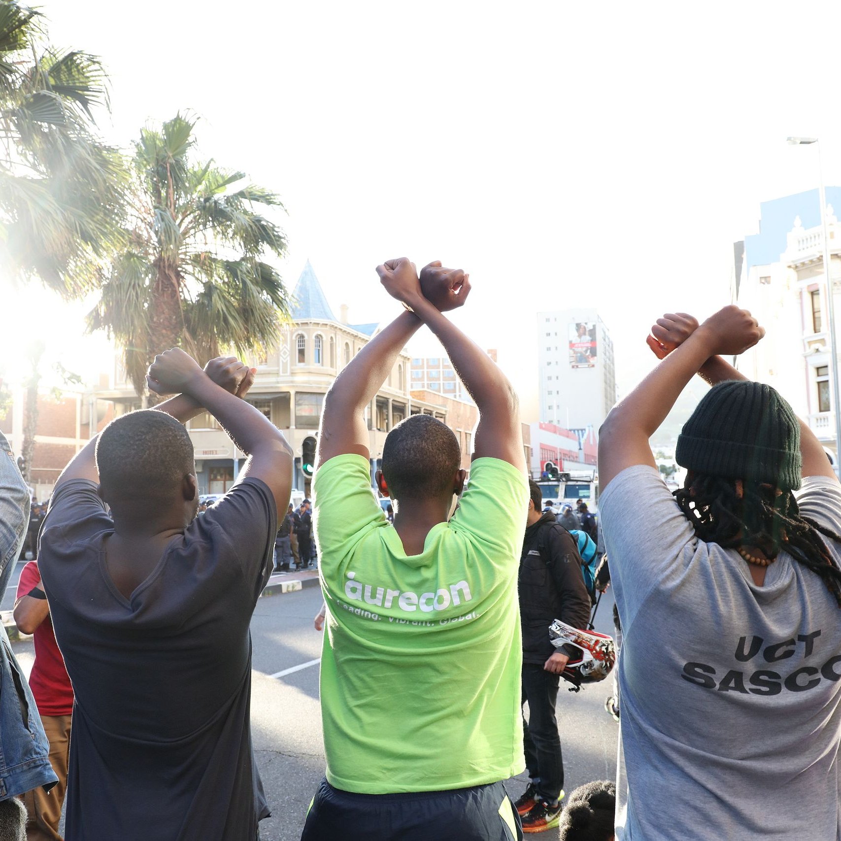 L'image montre un groupe de personnes vues de dos, levant les bras au-dessus de leurs têtes, formant un geste de solidarité ou de protestation. Ils sont sur une rue animée, entourés de bâtiments urbains. On peut apercevoir des palmiers à l'arrière-plan, ce qui ajoute une note de chaleur à l'environnement. La lumière du soleil éclaire la scène, créant une ambiance dynamique. Les personnes portent des t-shirts de différentes couleurs, dont l'un est vert vif. L'ensemble dégage une impression de union et d'engagement collectif.