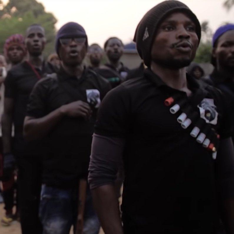L'image montre un groupe de jeunes hommes debout en formation. Ils portent des t-shirts noirs et semblent participer à une activité collective, probablement un chant ou un cri de ralliement. Certains d'entre eux tiennent des bâtons ou des armes. En arrière-plan, on aperçoit d'autres personnes rassemblées, avec des arbres et un paysage rural. L'atmosphère semble être à la fois solennelle et engagée. La lumière du soir crée une ambiance particulière. 