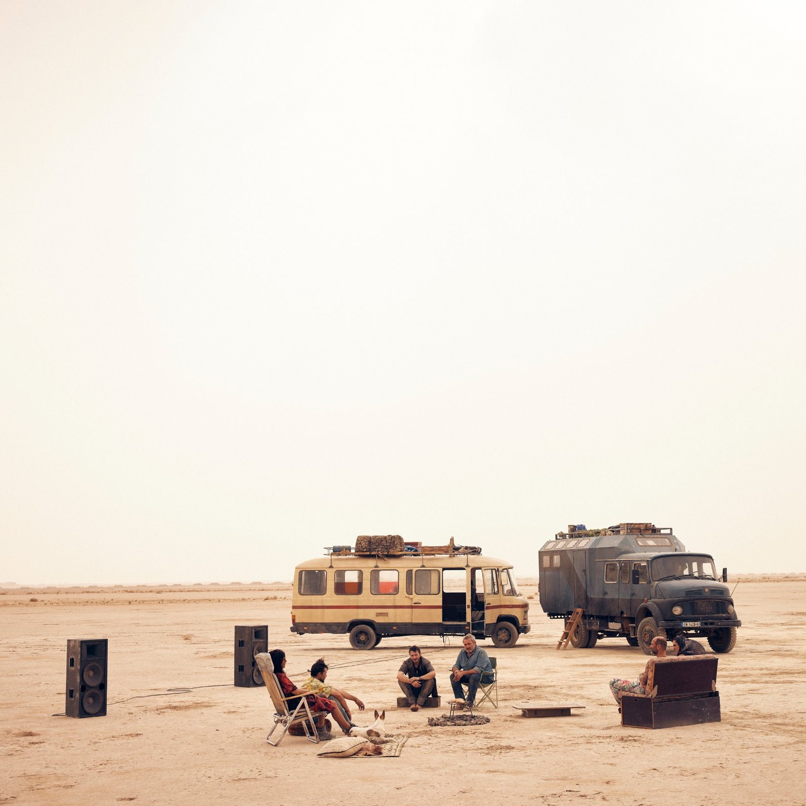 L'image montre un paysage désertique, vaste et aride, baigné par une lumière douce. Au centre, un groupe de personnes est assis autour d'un feu de camp. Ils semblent se détendre et passer un bon moment ensemble. À proximité, deux véhicules distincts attirent l'attention : l'un est un camping-car jaune, l'autre un véhicule utilitaire de couleur sombre. Des haut-parleurs ont été disposés autour du groupe, évoquant une ambiance festive. Le sol est sablonneux, et l'atmosphère semble calme, presque magique, sous un ciel d'une teinte uniforme.