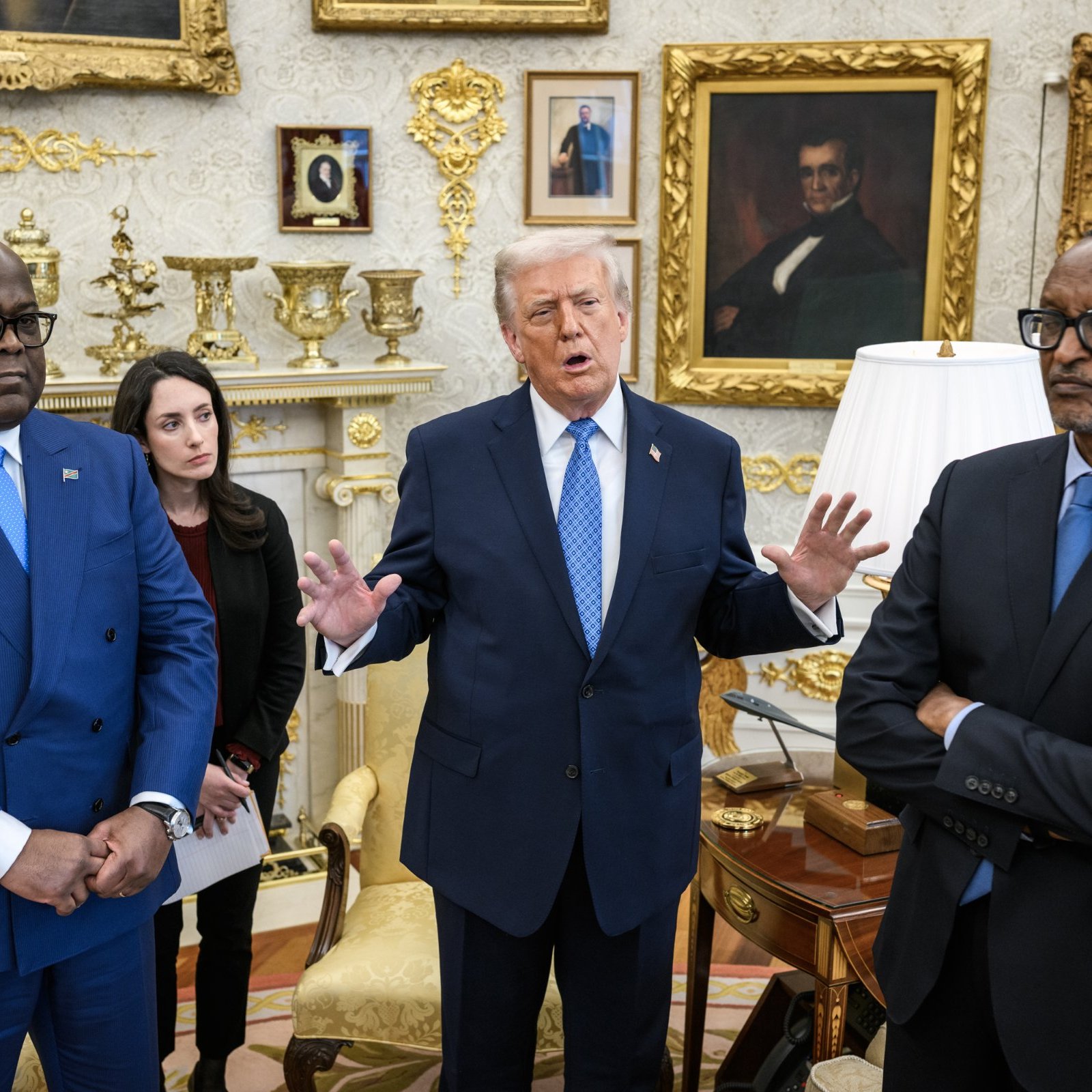 Dans cette image, nous sommes dans le Bureau Ovale, un espace formel et décoré richement. Au centre, un homme, probablement une figure politique, gesticule en s'exprimant avec animation. À sa gauche, un homme en costume bleu écoute attentivement, tandis qu'à droite, un autre homme, également en costume sombre, semble plus réservé, les bras croisés. Une femme en arrière-plan prend des notes, portant un vêtement noir. L'ambiance de la scène montre des échanges significatifs, probablement liés à des discussions politiques ou diplomatiques. Les murs sont ornés de cadres dorés, avec des portraits et des éléments décoratifs qui ajoutent à l'élégance de la pièce.
