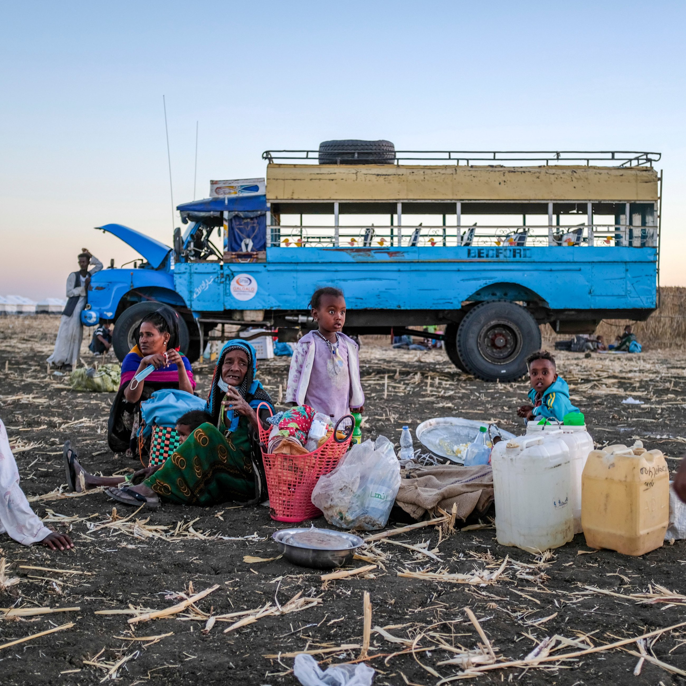 L'image montre un groupe de personnes assises sur un sol de terre battue, entourées de paniers et de contenants. Au centre, des femmes et des enfants semblent converser, tandis qu'un enfant se tient à droite, s'approchant. En arrière-plan, on peut voir un bus bleu immobile, avec un ciel dégradé qui passe des teintes orangées aux bleues, suggérant un crépuscule. Le terrain est jonché de restes de culture, ajoutant un contexte rural à la scène. L'atmosphère évoque une ambiance de rassemblement communautaire dans un environnement de travail.