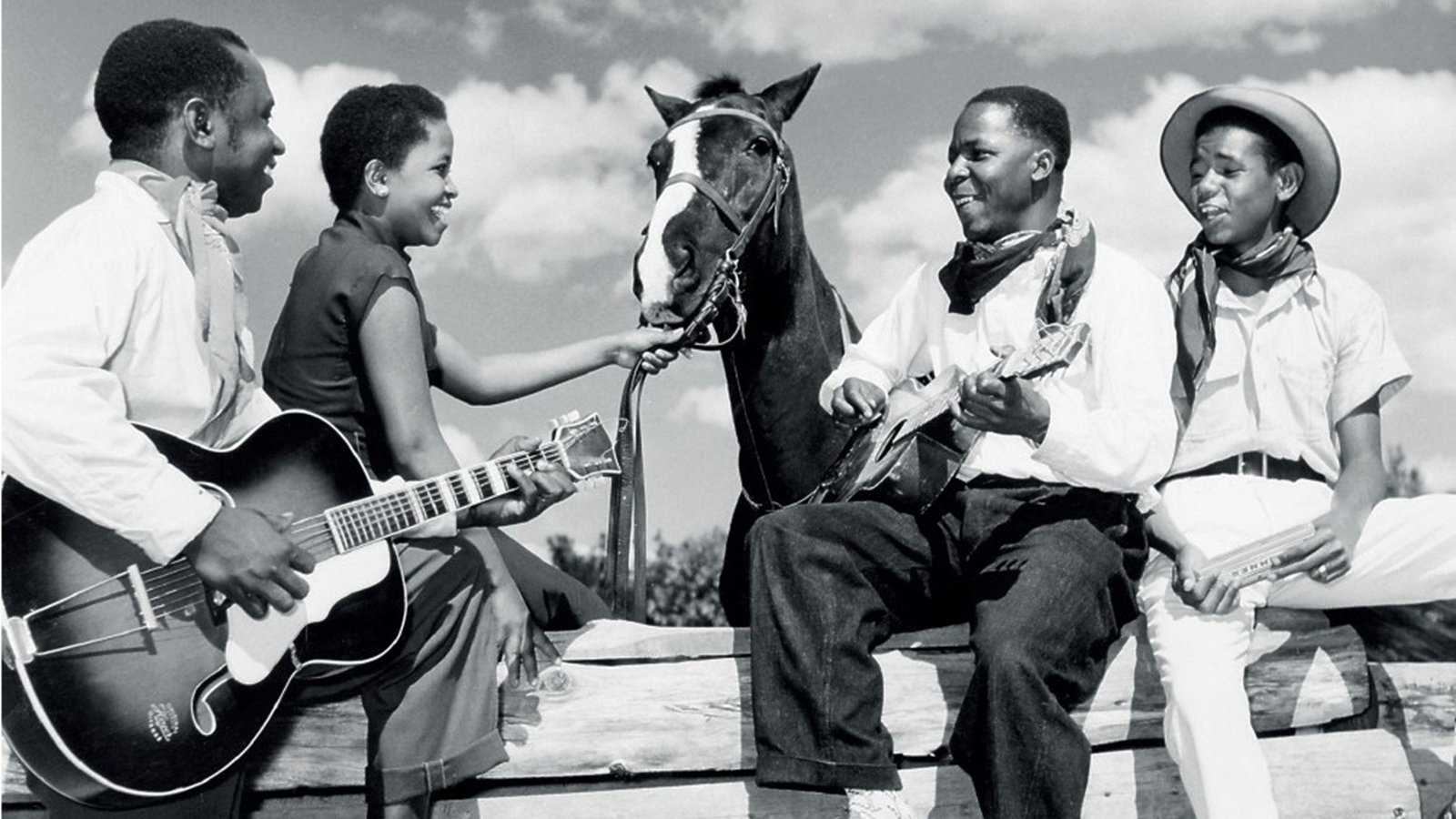 L'image représente un groupe de quatre personnes en noir et blanc, assises autour d'une barrière en bois, sous un ciel partiellement nuageux. Deux hommes et deux femmes sont présents. L'un des hommes joue de la guitare, tandis que l'autre est assis sur un tronc, tenant un instrument à cordes, probablement un banjo ou un ukulélé. Les deux femmes semblent interagir avec l'un des hommes et un cheval qui est également présent, la tête tournée vers eux. Elles affichent des expressions joyeuses, créant une atmosphère conviviale et détendue. Les vêtements des personnages varient : l'un porte un chapeau de cowboy et les autres portent des vêtements simples, typiques de l'époque. L'image évoque un moment de camaraderie et de musique, suggérant un lien fort entre les personnes et la nature environnante.
