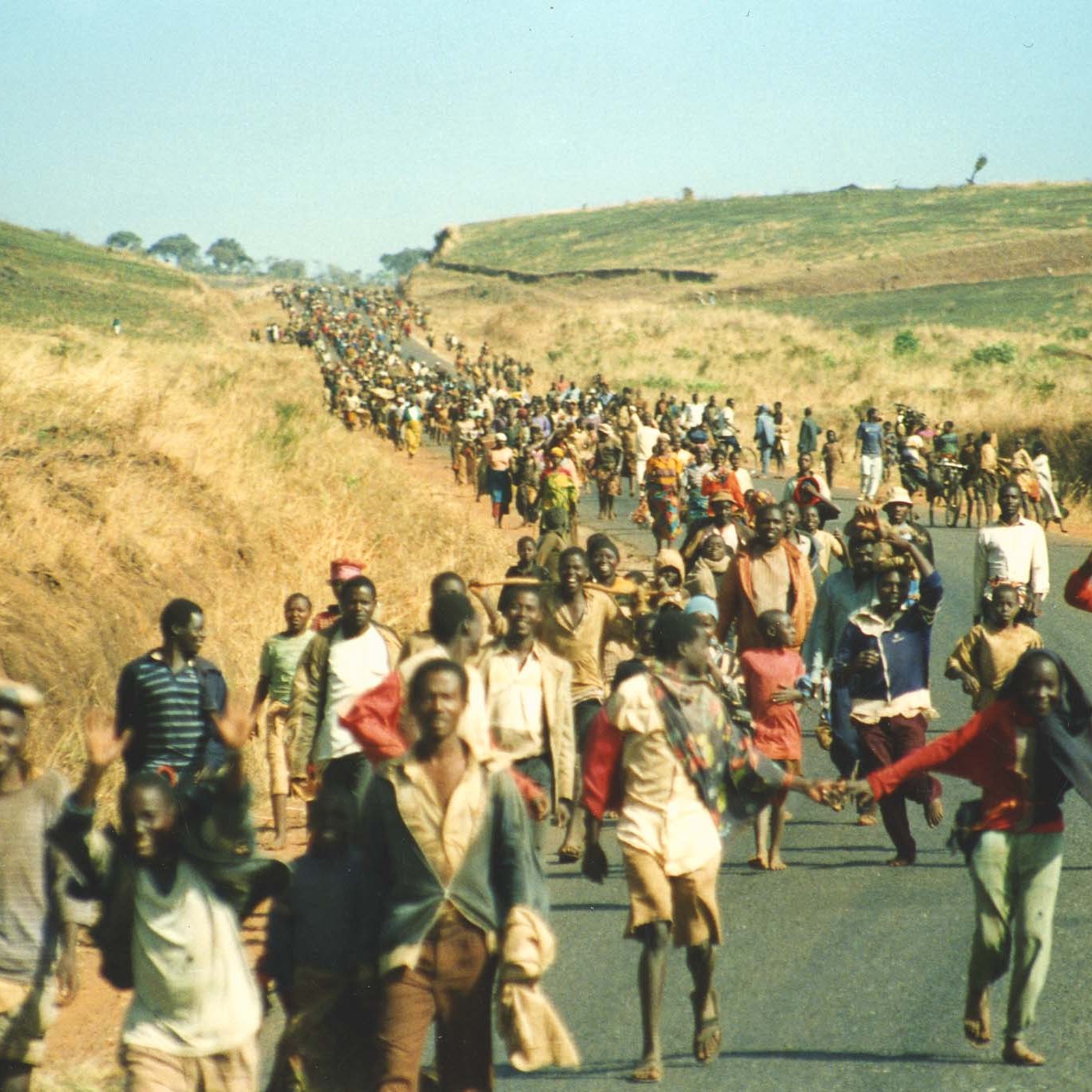 L'image montre une vaste foule de personnes marchant le long d'une route au milieu d'un paysage de collines sèches et herbeuses. Les individus, de différents âges et costumes, avancent ensemble, certains se tenant par la main ou portant des charges. L'atmosphère semble à la fois déterminée et solennelle, peut-être symbolisant un déplacement ou une migration collective. En arrière-plan, on aperçoit une étendue de terre vallonnée qui s'étend à perte de vue, ajoutant une dimension de profondeur à la scène. Le ciel est dégagé, laissant entrevoir un jour ensoleillé.