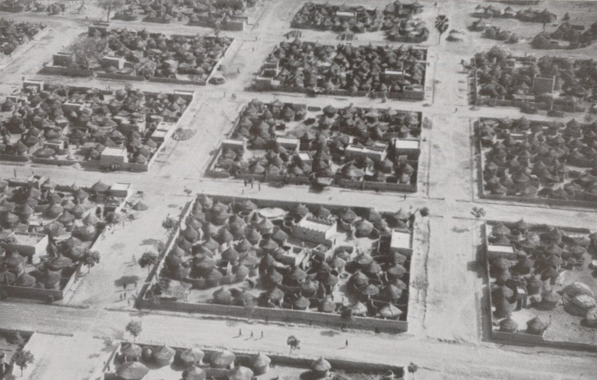 L'image montre une vue aérienne d'un village africain. On peut voir un agencement organisé des habitations, qui sont principalement des maisons traditionnelles en toit de chaume, caractéristiques de certaines régions. Les structures sont regroupées en plusieurs blocs délimités par des chemins. Des arbres sont dispersés dans l'espace, ajoutant une touche de verdure. Quelques personnes se déplacent le long des routes, suggérant une animation dans ce décor rural. Le photographique est en noir et blanc, ce qui donne une atmosphère historique à la scène.