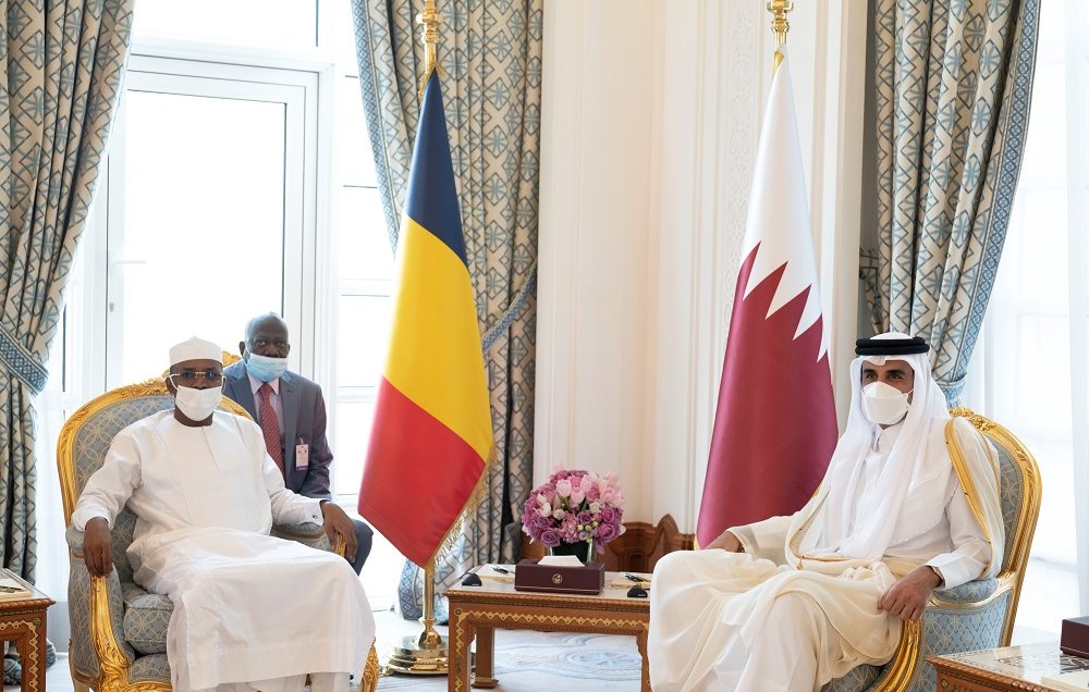 L'image montre une réunion formelle entre deux dirigeants. À gauche, un homme porte une tenue traditionnelle blanche et un chapeau, symbole de son pays. À sa droite, un autre homme est habillé d'une robe longue beige et d'un couvre-chef traditionnel. Entre eux, une table basse en bois contient des objets décoratifs. En arrière-plan, on peut voir des drapeaux : celui du Tchad à gauche et celui du Qatar à droite. La décoration des murs et des rideaux est élégante, avec des motifs riches et des couleurs apaisantes. L'atmosphère est solennelle et professionnelle.