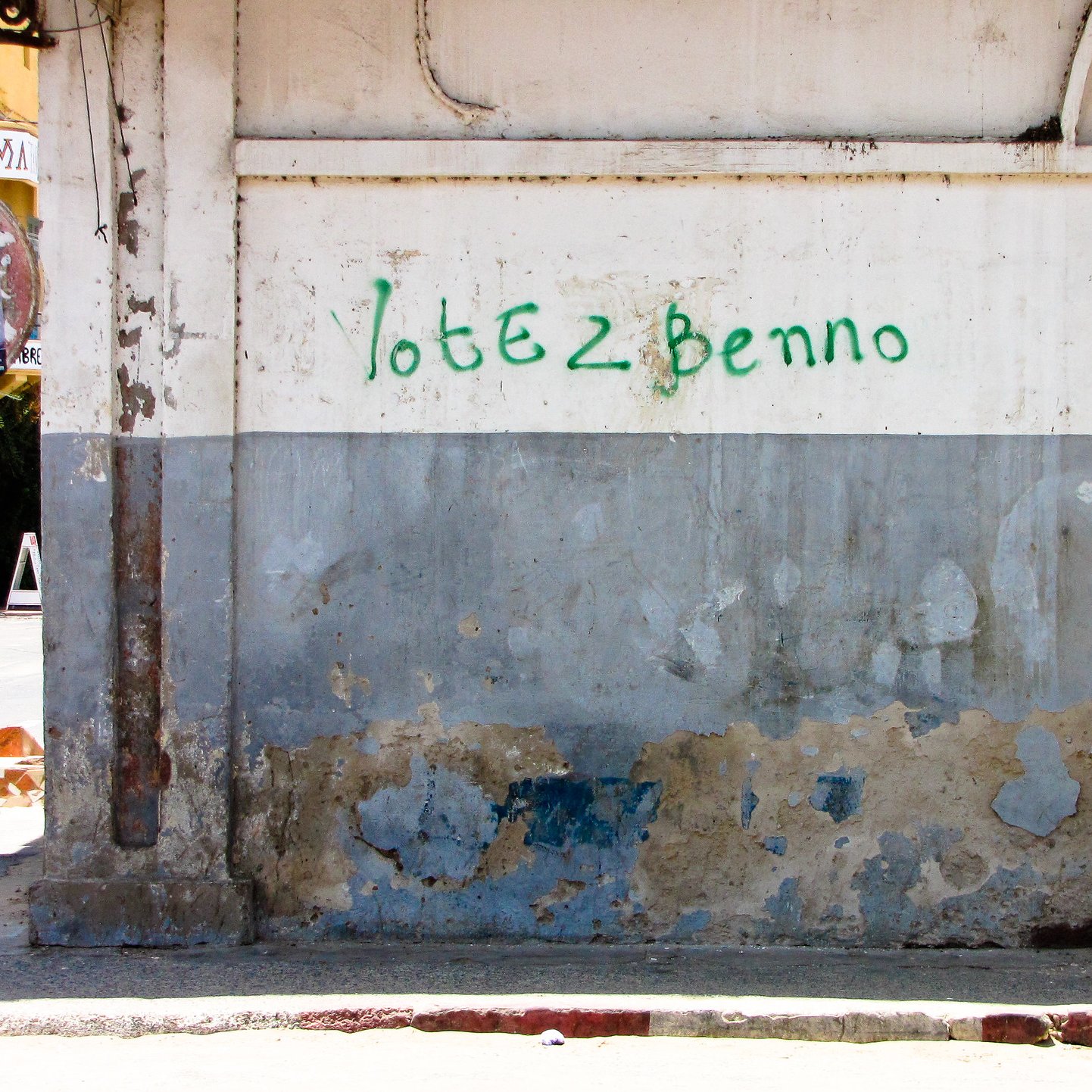 Cette image montre un mur extérieur dans une rue urbaine. La surface du mur est usée, avec de la peinture écaillée et des marques d'usure qui révèlent le temps qui passe. Sur la partie supérieure du mur, il y a une inscription verte qui dit "Votez Benno". À côté du mur, un homme est visible de dos, appuyé contre le mur. Son style vestimentaire semble décontracté. En arrière-plan, on peut apercevoir un peu de végétation et d'autres bâtiments qui enrichissent le paysage urbain. L'atmosphère générale évoque une scène de la vie quotidienne dans une ville animée.