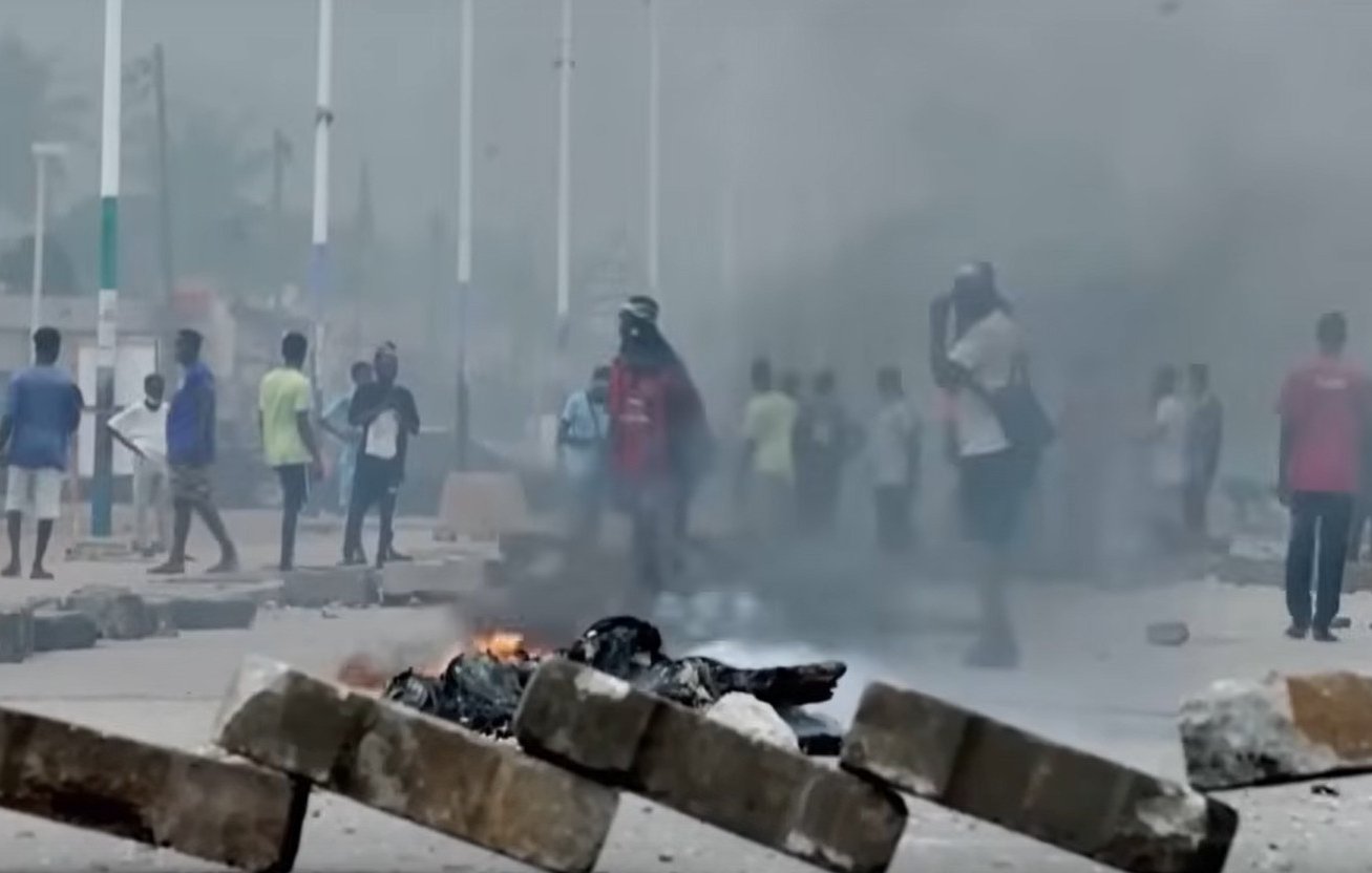 L'image montre une scène urbaine troublée. Au premier plan, plusieurs personnes courent rapidement, manifestant une impression d'urgence et de panique. En arrière-plan, de grands nuages de fumée noire s'élèvent dans l'air, indiquant un incendie. Des flammes orange et rouges jaillissent de barricades, suggérant une situation de conflit ou de désordre. Le décor urbain autour d'eux est flou, mais on peut discerner des bâtiments et des routes. L'atmosphère est tendue, évoquant un sentiment de danger et d'agitation.