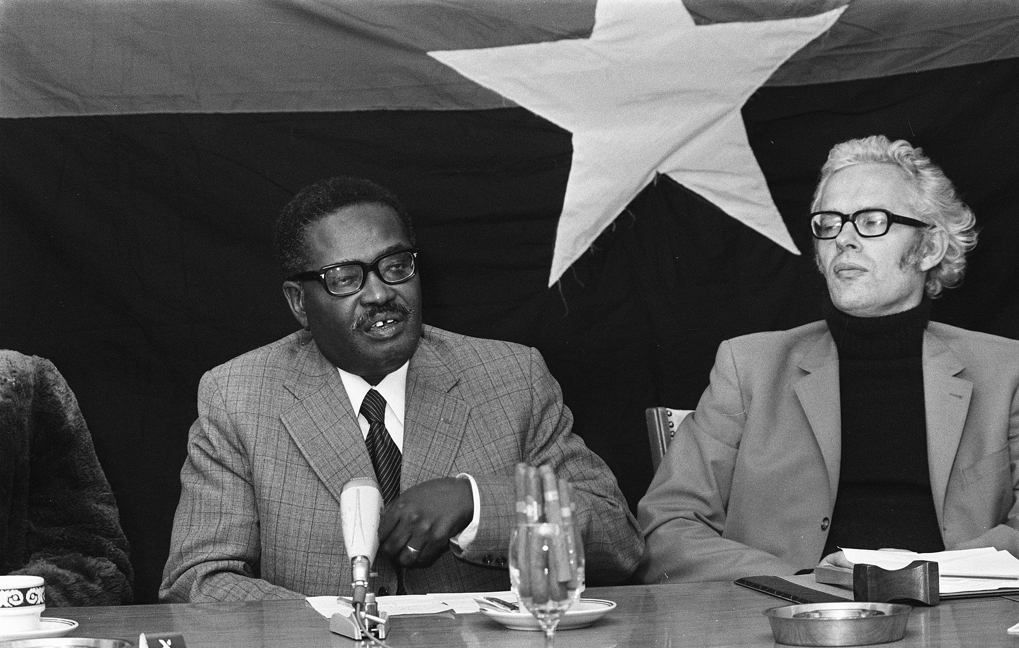 L'image montre une réunion où deux hommes sont assis à une table. À gauche, un homme d'âge moyen, portant des lunettes et un costume gris à carreaux, semble s'adresser aux autres participants, gesticulant légèrement avec une main. Il a une expression attentive et engagée. À sa droite, un homme plus âgé avec des cheveux gris et des lunettes, habillé d'un pull noir et d'une veste beige, l'écoute avec intérêt. En arrière-plan, on devine un grand drapeau noir avec une étoile blanche, ajoutant un contexte politique à la scène. Sur la table, il y a quelques verres, un microphone et un bloc-notes. L'atmosphère semble sérieuse et concentrée.