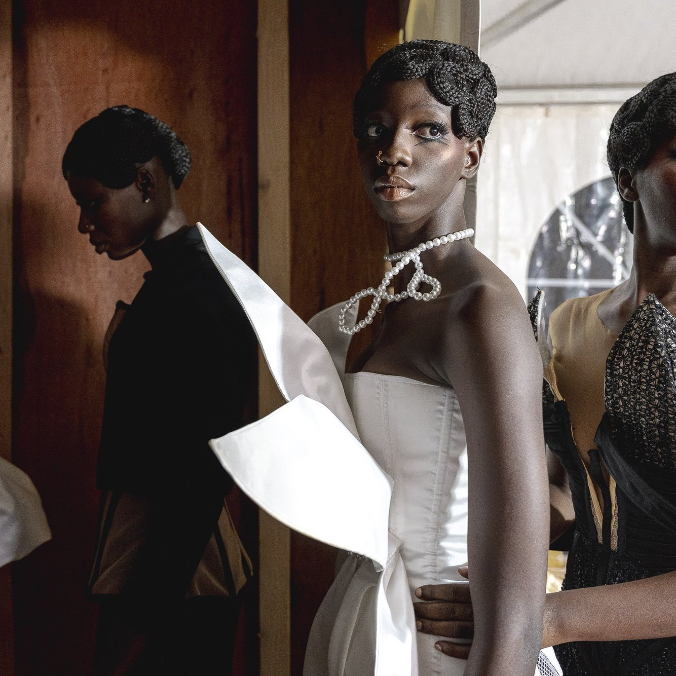 L'image présente un groupe de mannequins lors d'un événement de mode. Les mannequins, d'origines diverses, portent des tenues élégantes et extravagantes, mettant en valeur des tissus texturés et des coupes audacieuses. Au premier plan, l'un des mannequins se distingue par une robe blanche ornée d'un grand nœud, tandis que les autres vêtus de créations plus sombres, affichent des coiffures soignées. La lumière joue sur les tissus, créant un contraste entre les ombres et les zones éclairées, ce qui ajoute une profondeur visuelle à la scène. L'ambiance est à la fois chic et artistique, capturant l'énergie dynamique du monde de la mode.