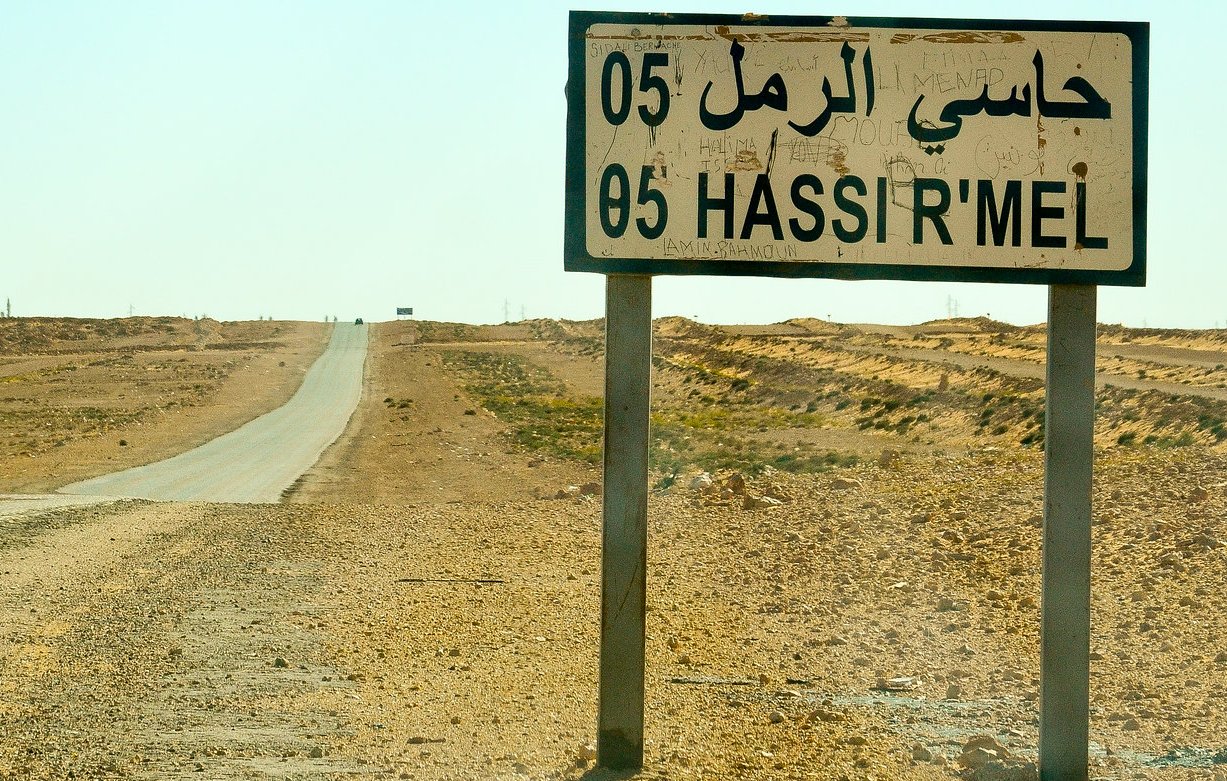 L'image montre une route désertique dans un paysage aride. À droite, un grand panneau de signalisation indique "Hassi R'Mel" en arabe et en français, avec le numéro 05. En arrière-plan, on aperçoit des structures industrielles, dont une cheminée qui dégage de la fumée, suggérant l'activité d'extraction de ressources. L'environnement est principalement constitué de terres sèches et peu de végétation, illustrant une atmosphère isolée et stérile. Le ciel est clair, et la chaleur du jour est palpable.