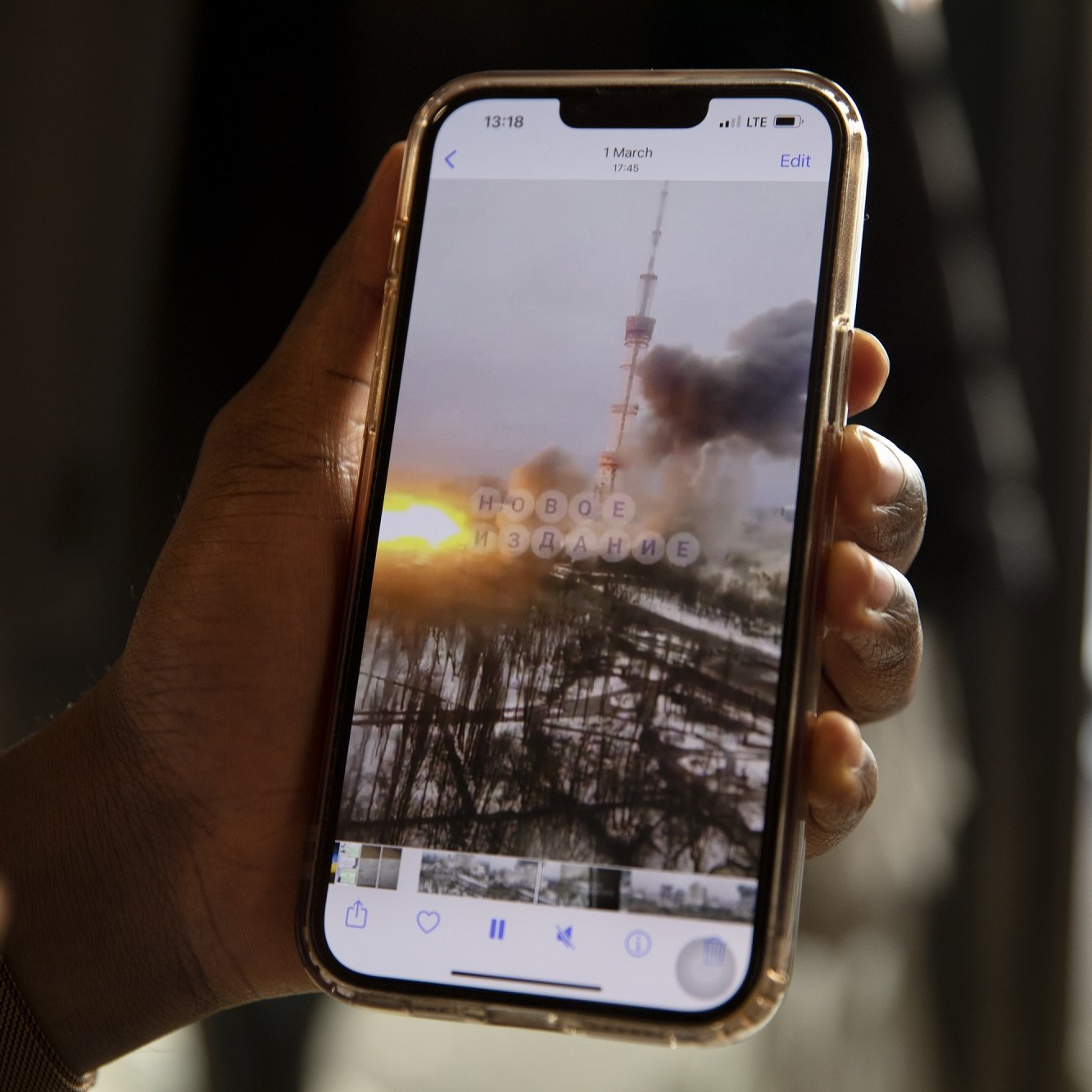 L'image montre une main tenant un smartphone dans un environnement intérieur. Sur l'écran du téléphone, on peut voir une photo qui semble représenter un paysage urbain, où une tour se dresse au milieu de colonnes de fumée et des nuages sombres. À l'arrière-plan, un coucher de soleil offre des teintes orange et jaune qui contrastent avec la grisaille de la scène. Les détails flous et éthérés suggèrent une atmosphère dramatique, peut-être liée à un événement perturbant. La main qui tient le téléphone est d'une peau foncée et porte un bracelet lumineux, ajoutant une touche personnelle à l'image.
