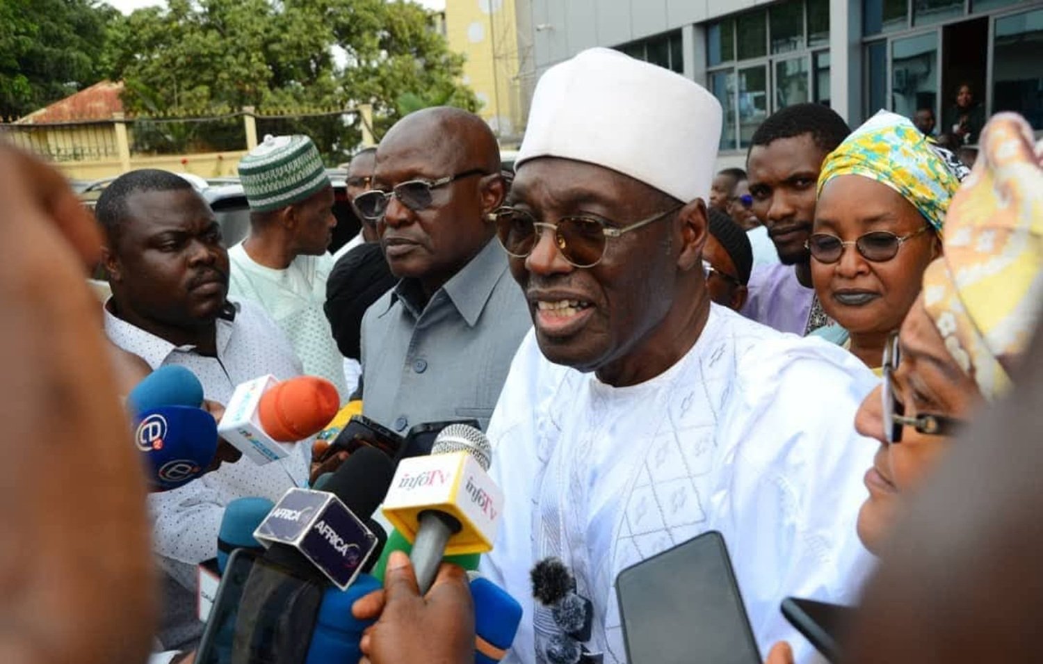 The image shows a group of people gathered around a man wearing a white traditional outfit and a white cap. He appears to be speaking to the media, as indicated by the microphones and cameras directed at him. Surrounding him are several other individuals, some of whom are dressed in formal attire, while others wear traditional garments. The background features a building, and the atmosphere suggests a press event or a public announcement.