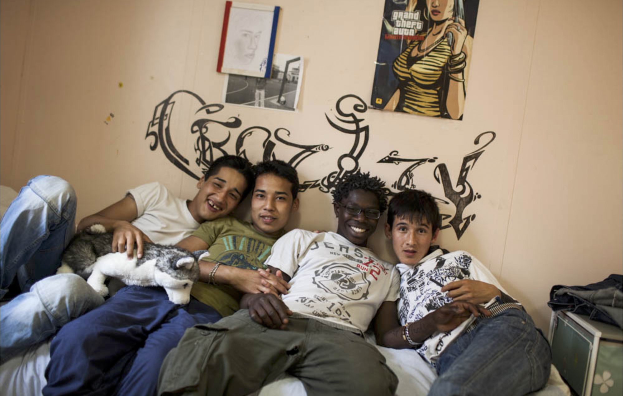 L'image montre quatre jeunes hommes posant ensemble sur un lit dans une chambre. Ils sourient, affichant une ambiance amicale et détendue. La pièce est décorée simplement, avec un mur où l'on peut voir des graffitis. À côté d'eux, un petit chat est blotti. L'un des jeunes tient un ours en peluche. Chaque personne présente une tenue décontractée. Les expressions sur leurs visages suggèrent un moment de complicité et de camaraderie.