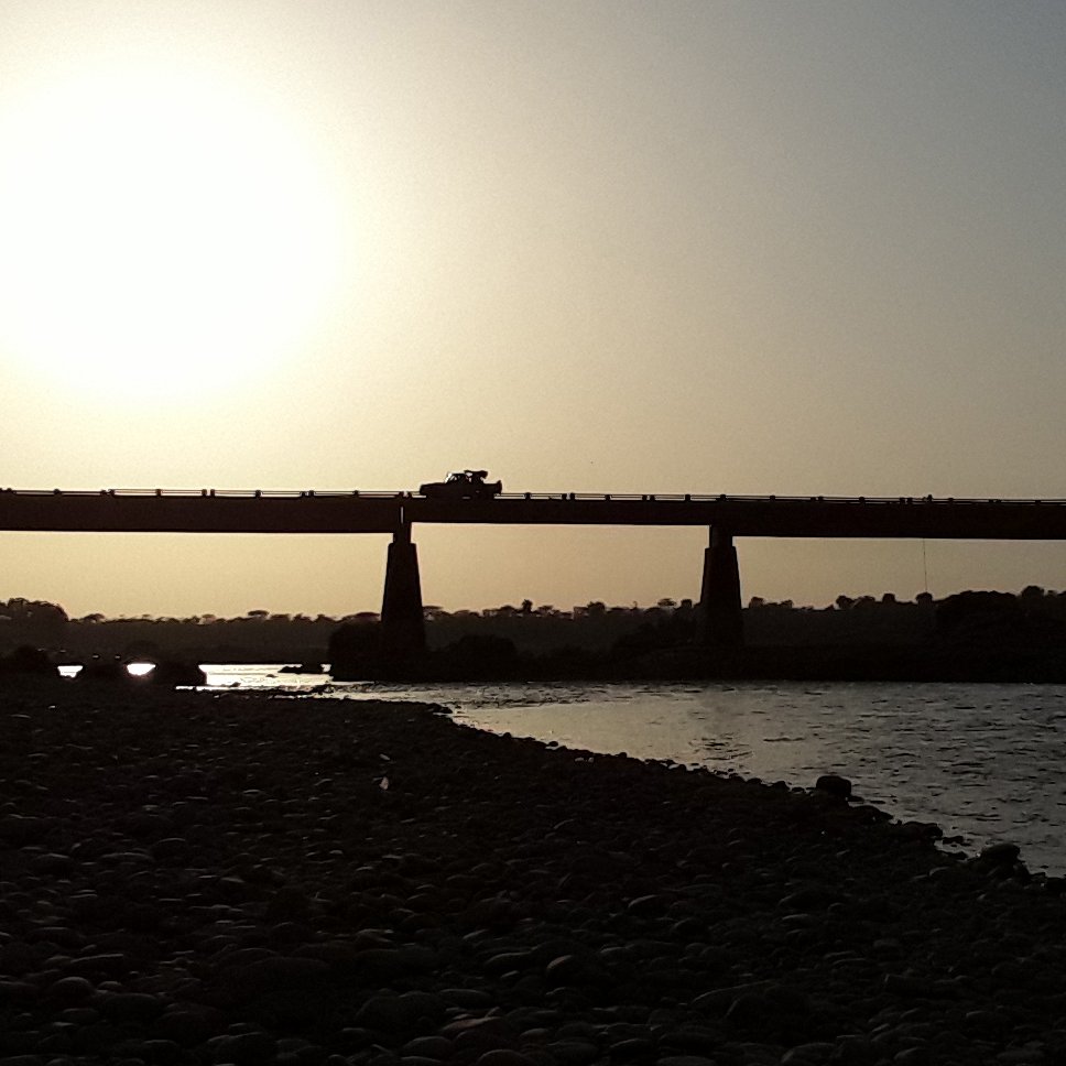 L'image montre un paysage côtier au crépuscule. À l'arrière-plan, on aperçoit un grand pont qui s'étend au-dessus d'une rivière. La lumière du soleil, basse à l'horizon, crée un contre-jour spectaculaire, rendant le pont et les éléments environnants en silhouette. Les contours de la structure sont sombres, se détachant des teintes dorées et oranges du ciel. À proximité de la rive, on peut imaginer des rochers ou des galets, ajoutant une texture au paysage. L'atmosphère est calme et paisible, évoquant un moment de tranquillité près de l'eau.