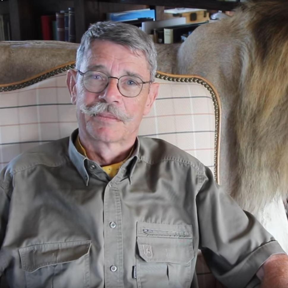 L'image montre un homme assis sur un fauteuil. Il a des lunettes, une moustache grise et porte une chemise kaki. À côté de lui, il y a un lion naturalisé, avec une crinière fournie et un regard majestueux. Le cadre donne une impression chaleureuse et rustique, avec une ambiance intérieure qui suggère une maison ou un chalet. Le contraste entre l'homme et le lion évoque une connexion avec la nature sauvage.
