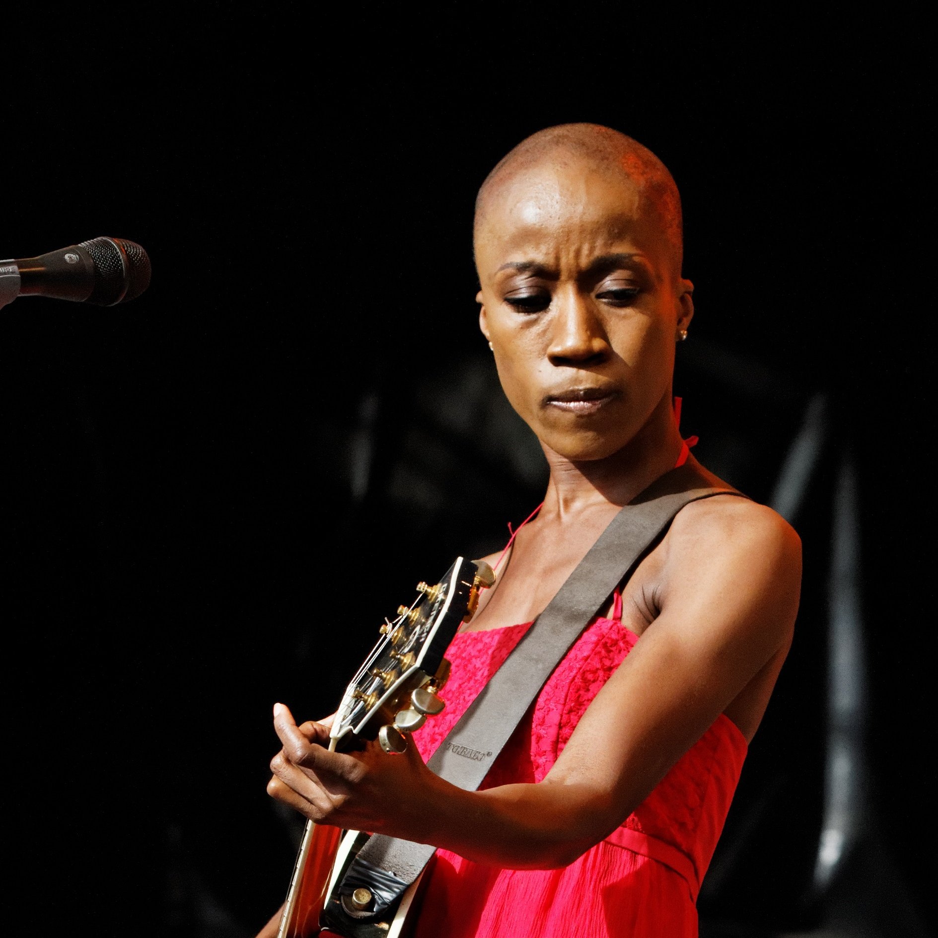 L'image montre une artiste sur scène qui joue de la guitare. Elle a la tête rasée, ce qui met en avant son visage expressif. Elle porte une robe rouge qui attire l'attention et contraste avec l'éclairage du fond sombre de la scène. Son regard est concentré, montrant sa passion et son engagement dans sa performance. Le microphone est placé devant elle, indiquant qu'elle chante probablement en même temps qu'elle joue. L'ambiance générale semble dynamique et remplie de musique, créant une atmosphère captivante.