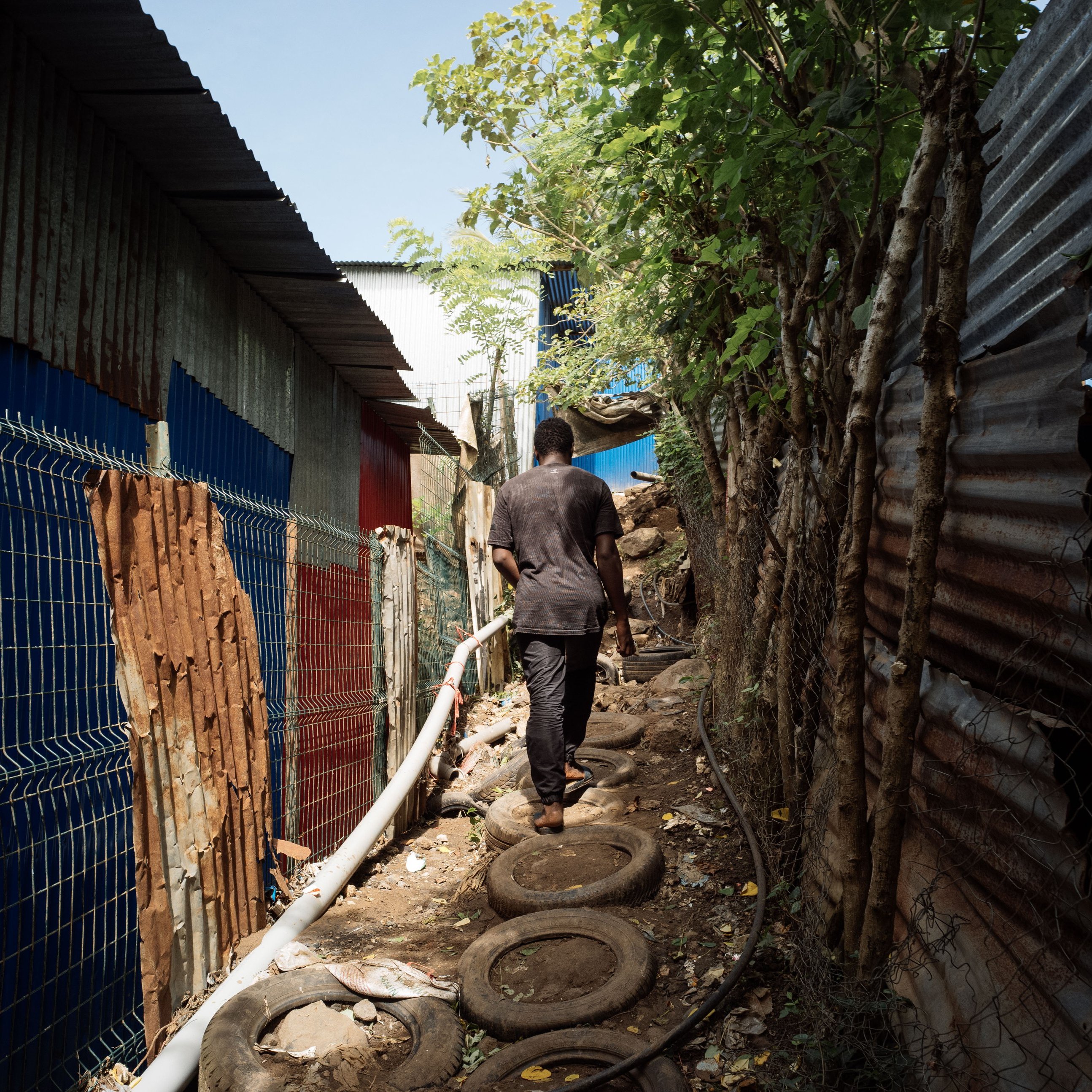 L'image montre un sentier étroit entre deux structures en tôle ondulée, typiques des bidonvilles. À gauche, on distingue des murs colorés, tandis qu'à droite, la structure est recouverte de tôles rouillées. Le chemin est constitué de pneus empilés, et un homme marche le long de ce chemin, portant des vêtements sombres. La lumière du jour illumine la scène, mettant en évidence la végétation qui borde le sentier, avec des arbres et des feuilles qui apportent une touche de verdure à cet environnement urbain. L'atmosphère semble à la fois animée et paisible.