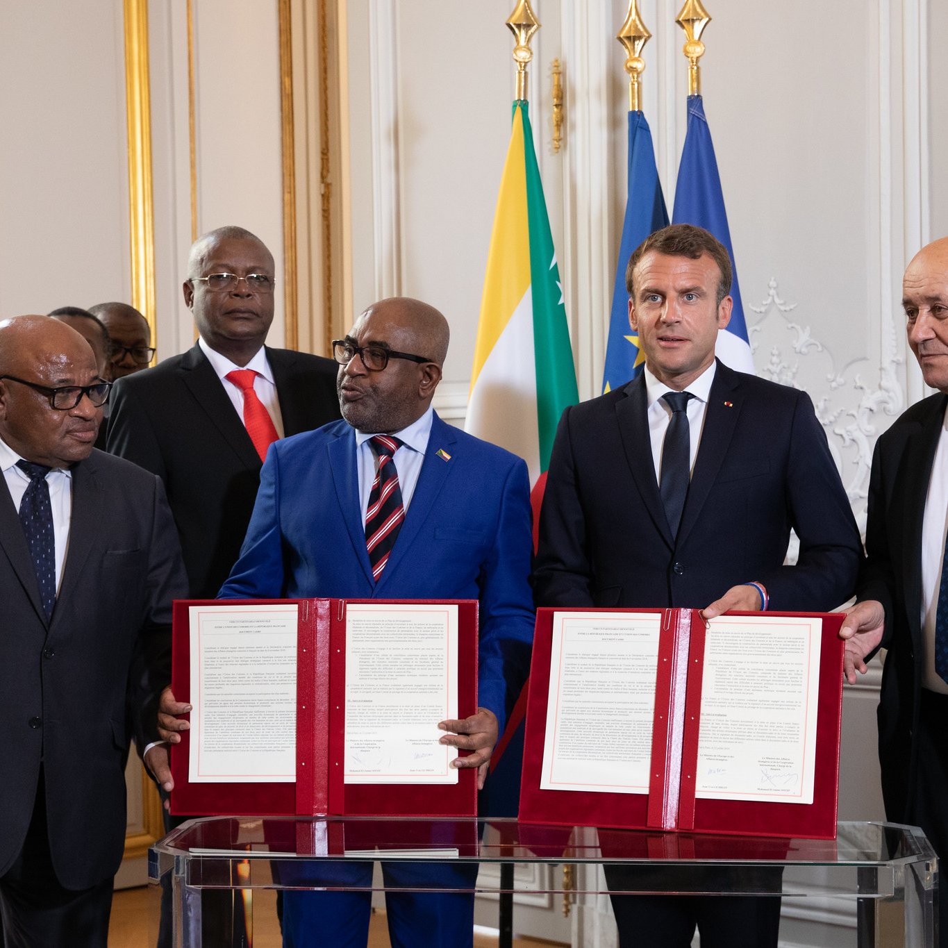 L'image montre un groupe de personnes lors d'une cérémonie officielle. Au centre, deux hommes tiennent des documents signés, symbolisant un accord ou un partenariat. L'un d'eux porte un costume bleu avec une cravate à motifs, tandis que l'autre est vêtu d'un costume sombre avec une cravate noir. En arrière-plan, on remarque des drapeaux, dont ceux de la France et d'autres pays africains. Les autres personnes en arrière-plan observant la scène portent également des costumes. L'ambiance générale est solennelle, mettant en avant un moment important de collaboration entre les nations.