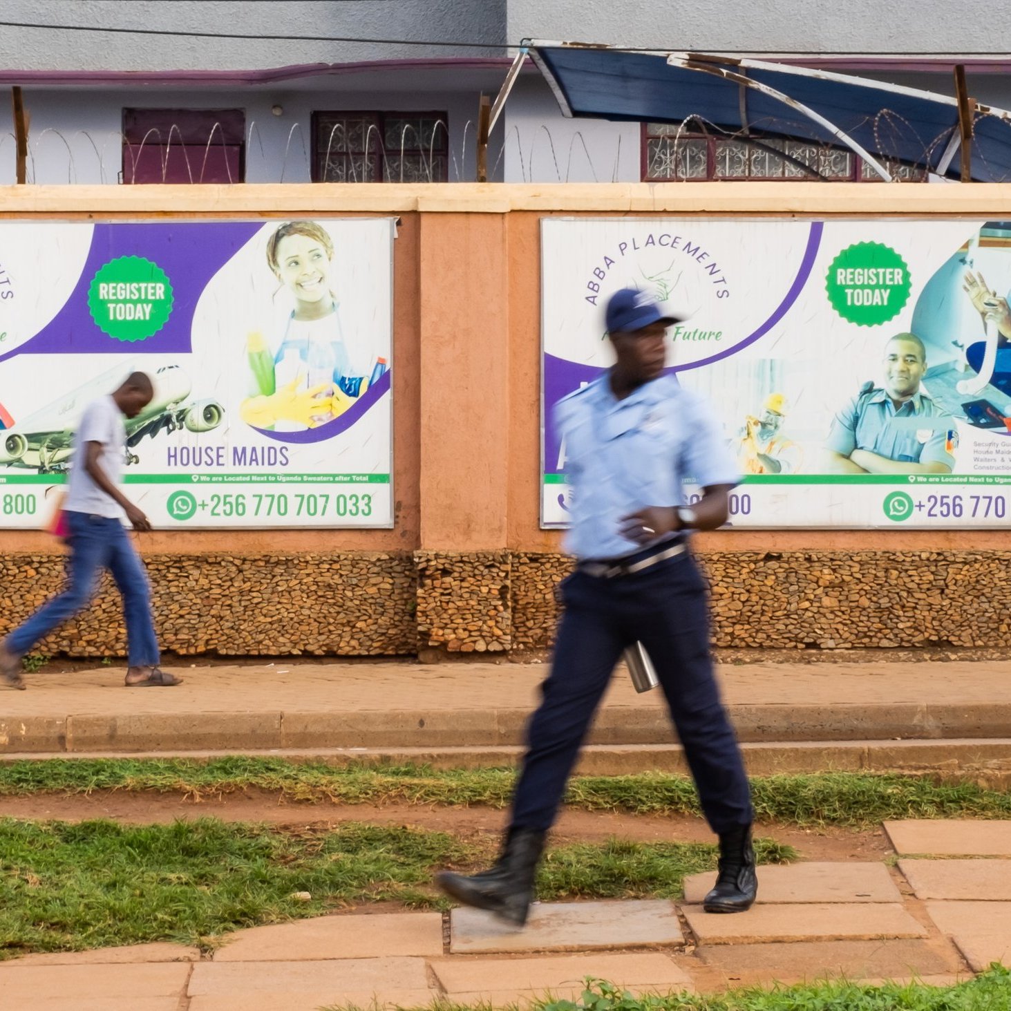L'image montre un environnement urbain animé. Sur un mur en bois, plusieurs affiches colorées sont accrochées, promouvant des services tels que des opportunités de travail à l'étranger et des services de ménage. Les affiches présentent des visages souriants de personnes, évoquant une atmosphère positive et encourageante. Au premier plan, un homme en uniforme, probablement un agent de sécurité, marche avec assurance sur le trottoir, tandis qu'un autre passant se déplace rapidement derrière lui. Le sol est pavé, et on peut deviner une verdure légère avec une pelouse bien entretenue. L'ensemble évoque une scène de la vie quotidienne dans une ville dynamique.