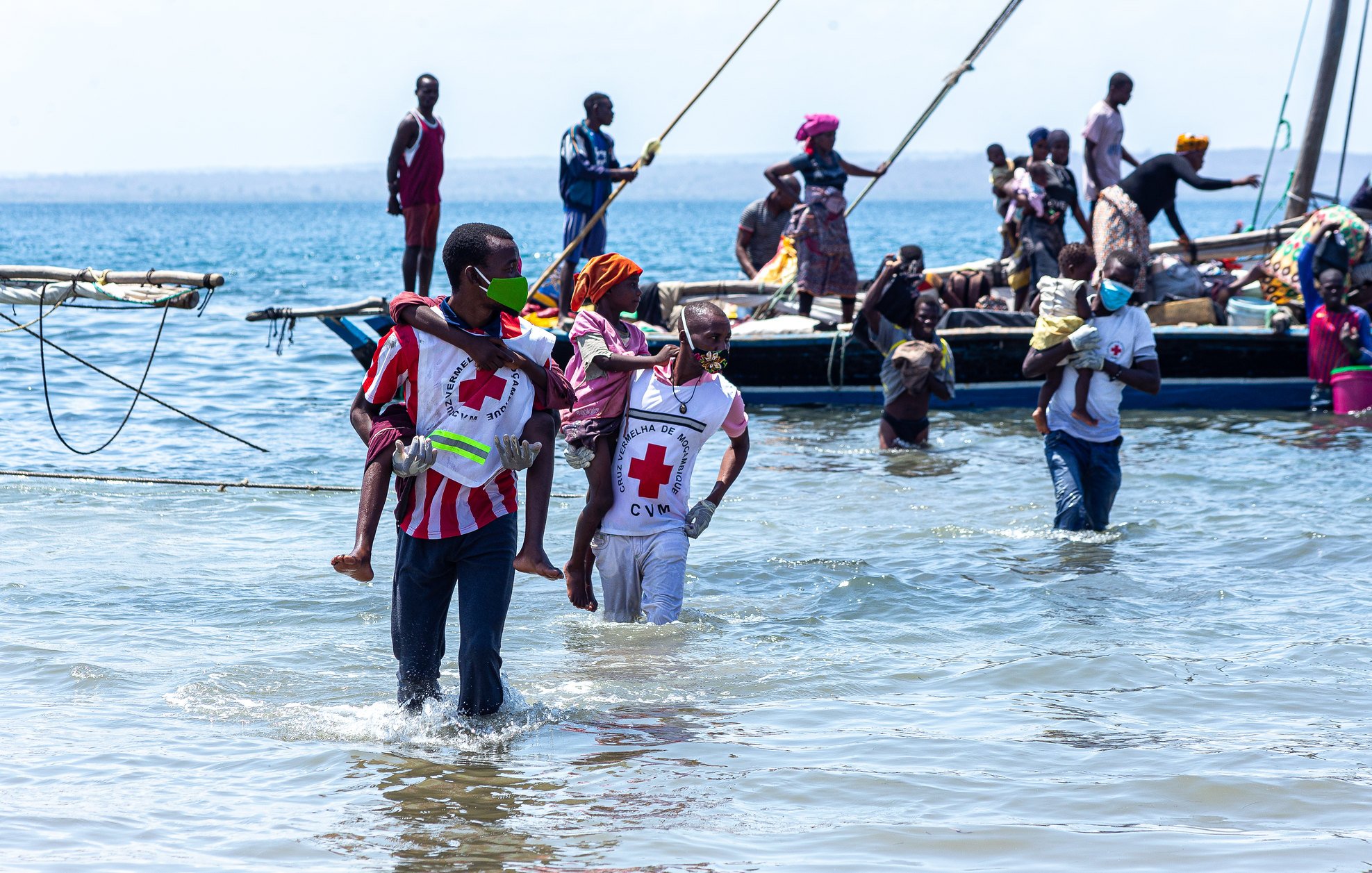L'image montre une scène animée au bord de la mer. On peut voir plusieurs personnes dans l'eau peu profonds, certaines portant des vêtements colorés. Des hommes et des femmes s'affairent autour de petites embarcations. Certaines personnes transportent des enfants, tandis que d'autres semblent s'entraider. En arrière-plan, il y a une barque avec des passagers qui attendent. L'ambiance semble être pleine de vie et de solidarité, avec un ciel dégagé et une mer calme.