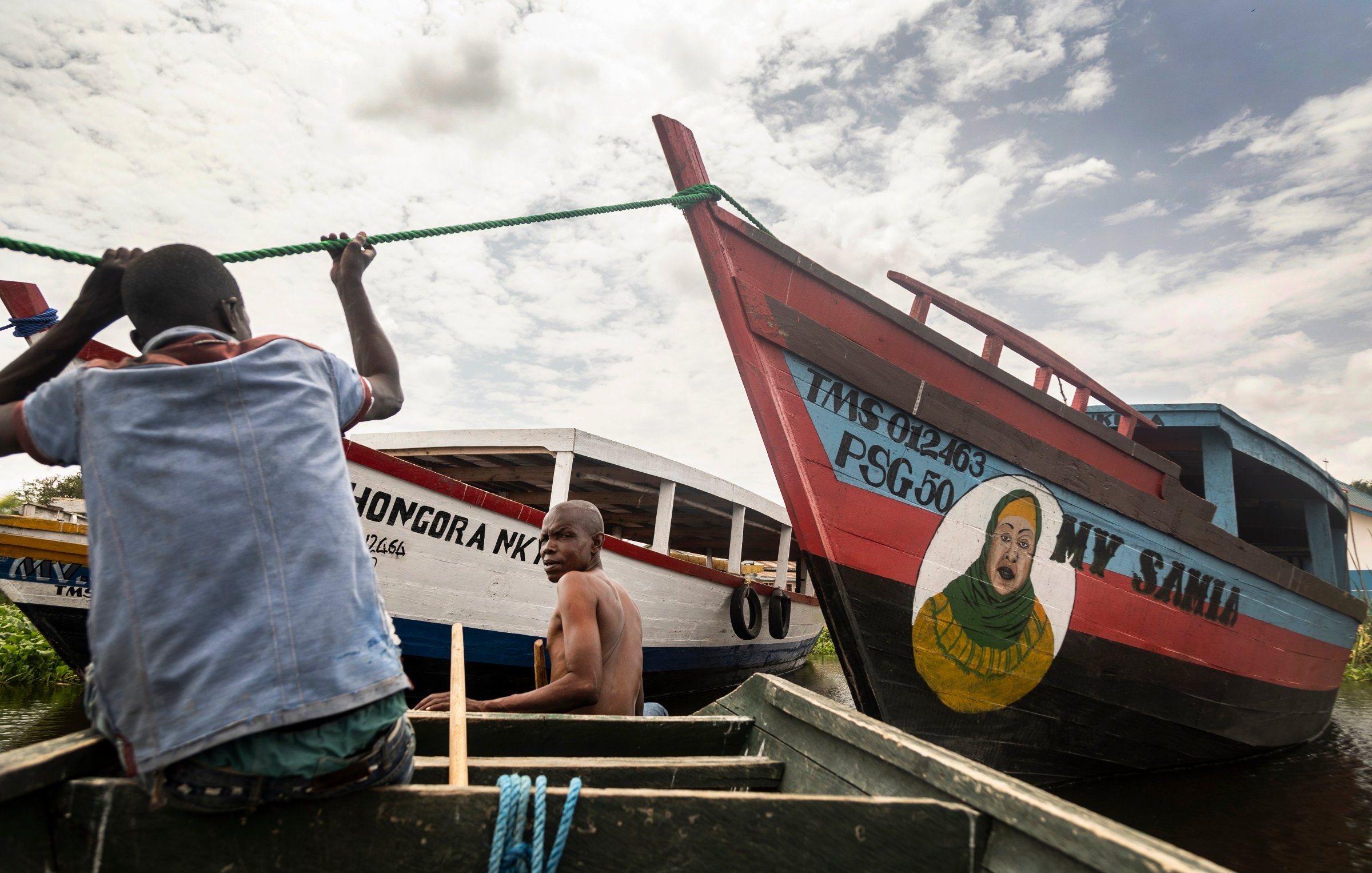L'image montre deux bateaux colorés amarrés au bord d'un cours d'eau calme. À l'avant, un homme, en train de ramer, est visible, concentré sur son effort. Un autre homme est assis à l'arrière, regardant vers l'objectif. Les bateaux sont peints de manière vive, avec des inscriptions comme "MY SAID" et "MONGORA MK". L'un des bateaux présente également une grande peinture d'un visage souriant. Le ciel est partiellement nuageux, ajoutant une dimension lumineuse à la scène. L'atmosphère est paisible, évoquant une ambiance de vie flottante.