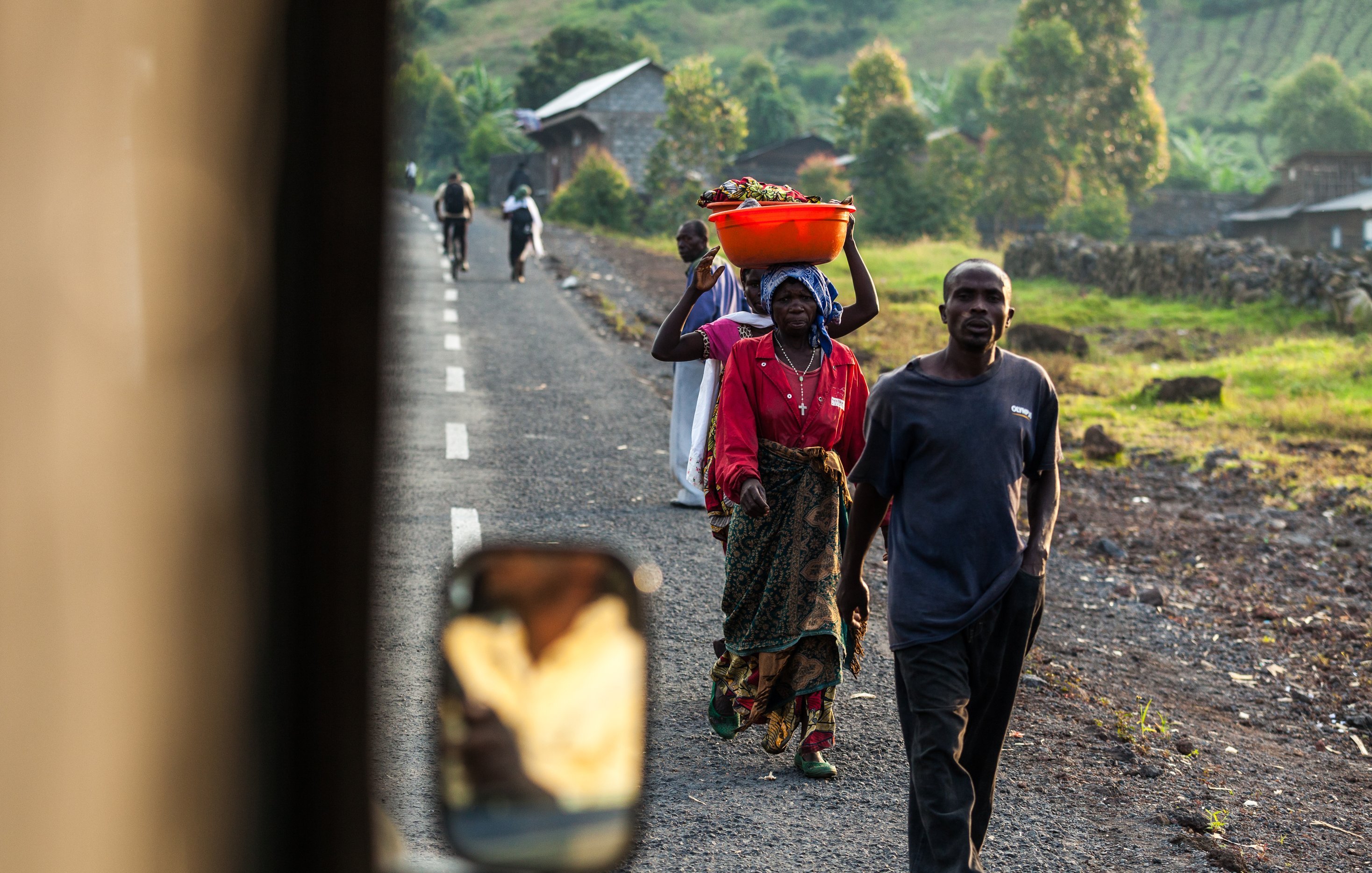 L'image montre une scène de vie quotidienne dans une région rurale. Sur une route en terre, plusieurs personnes marchent, chacune portant des charges sur leurs épaules ou dans les bras. Au premier plan, une femme est vue de dos, portant un grand panier orange rempli de fruits ou de légumes sur sa tête. Elle est vêtue d'une robe colorée. À côté d'elle, un homme porte une veste sombre et marche avec assurance. En arrière-plan, on peut apercevoir d'autres personnes se déplaçant sur la route, ainsi que des habitations et des collines verdoyantes qui donnent une impression de paysage naturel. La lumière du soleil éclaire la scène, créant une atmosphère chaleureuse et vivante.