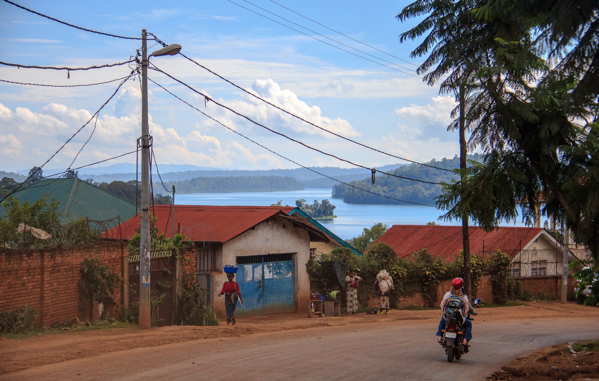 Voici une description de l'image : Vous vous trouvez sur une route située dans un paysage rural. À votre gauche, des maisons aux toits de tôle rouge et bleu se dressent, entourées de verdure luxuriante. Au loin, un lac scintille sous la lumière du soleil, reflétant les nuages blancs dans un ciel bleu. Certains habitants se déplacent, l'un d'eux portant une charge sur sa tête, tandis qu'un motard passe sur la route. Des poteaux électriques bordent la rue, ajoutant une touche de modernité à ce cadre naturel paisible. Le son lointain de l'eau et le chant des oiseaux peuvent également être présents dans cette ambiance tranquille.
