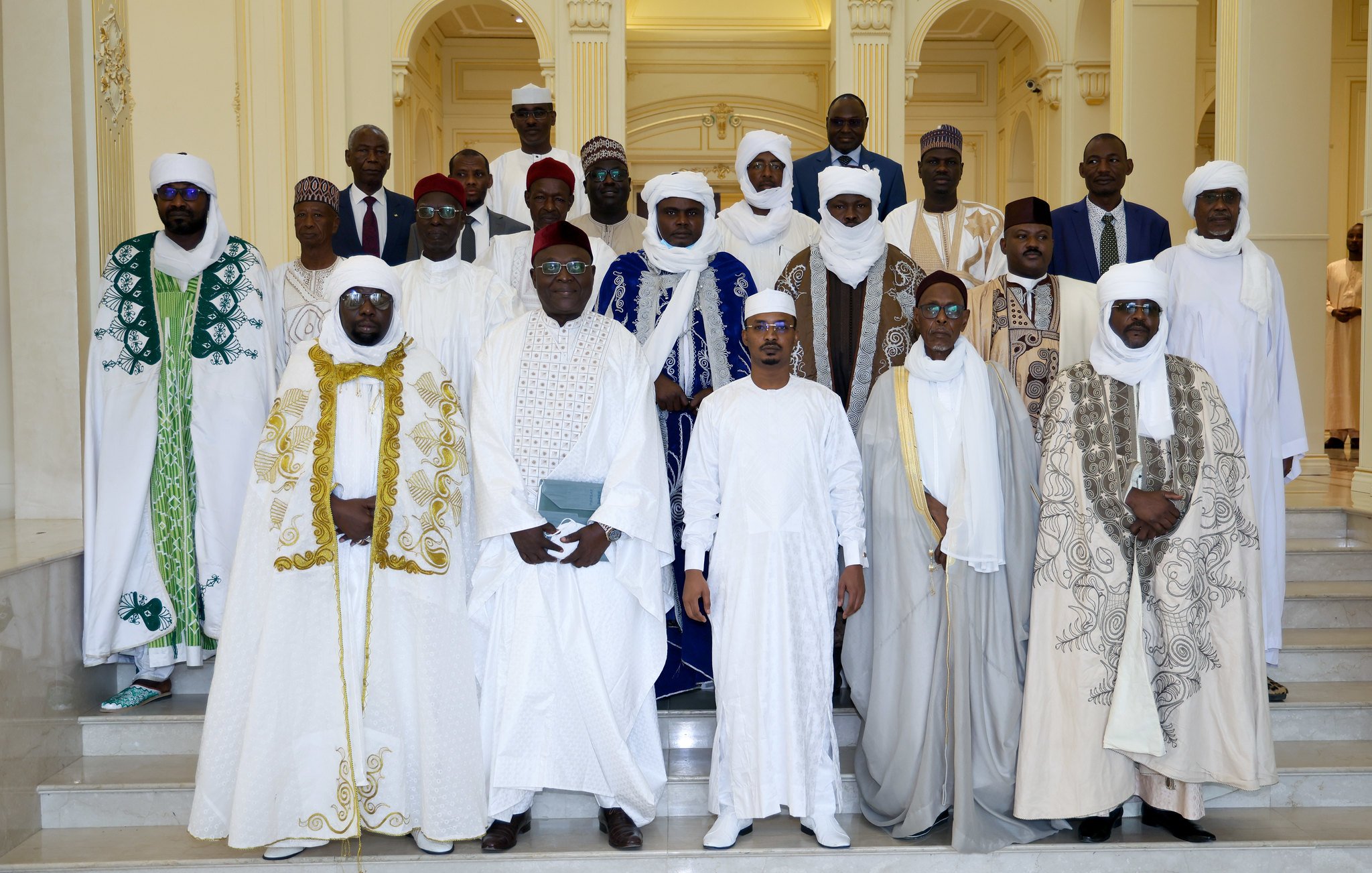 L'image montre un groupe de personnes habillées en vêtements traditionnels, rassemblées dans un espace élégant. Ils sont principalement vêtus de robes blanches, avec des éléments colorés comme des capes vertes et bleues. Les hommes portent des turbans et des chapeaux, ajoutant à la richesse de leurs tenues. L'arrière-plan est un intérieur lumineux et spacieux, avec des colonnes et des décorations architecturales. Le groupe semble coordonné et solennel, probablement pour une occasion spéciale.