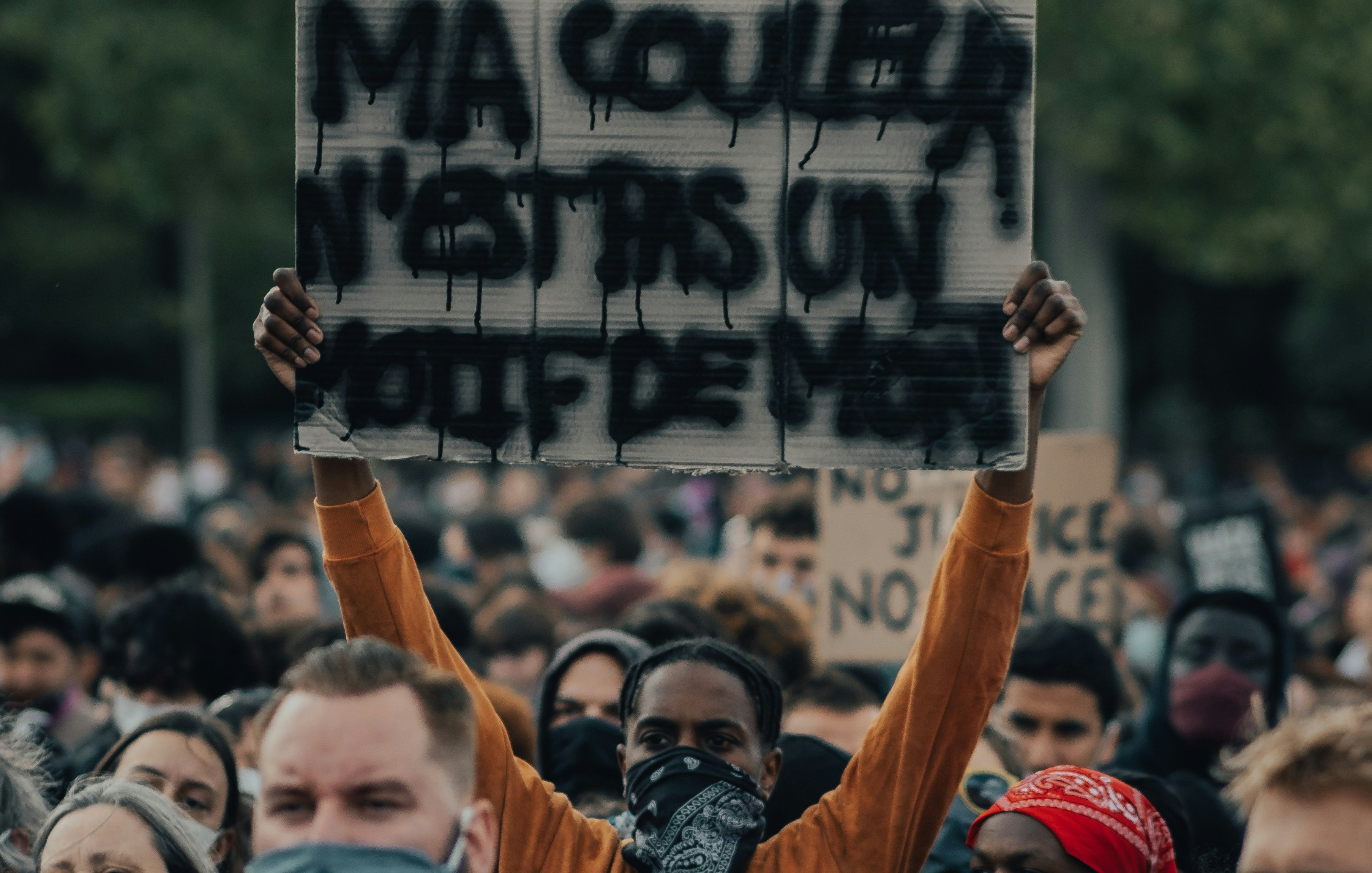 L'image montre une grande foule rassemblée pour une manifestation. Parmi les manifestants, une personne se distingue en tenant une pancarte sur laquelle est écrit "MA COULEUR N'EST PAS UN CRIME". Cette personne porte un masque, comme beaucoup d'autres dans la foule. Les visages autour sont sérieux et déterminés, reflétant un sentiment de solidarité et de revendication. On peut sentir l'intensité de l'événement grâce à l'énergie collective. En arrière-plan, des arbres indiquent un cadre naturel, ajoutant à l'atmosphère de mobilisation pacifique.