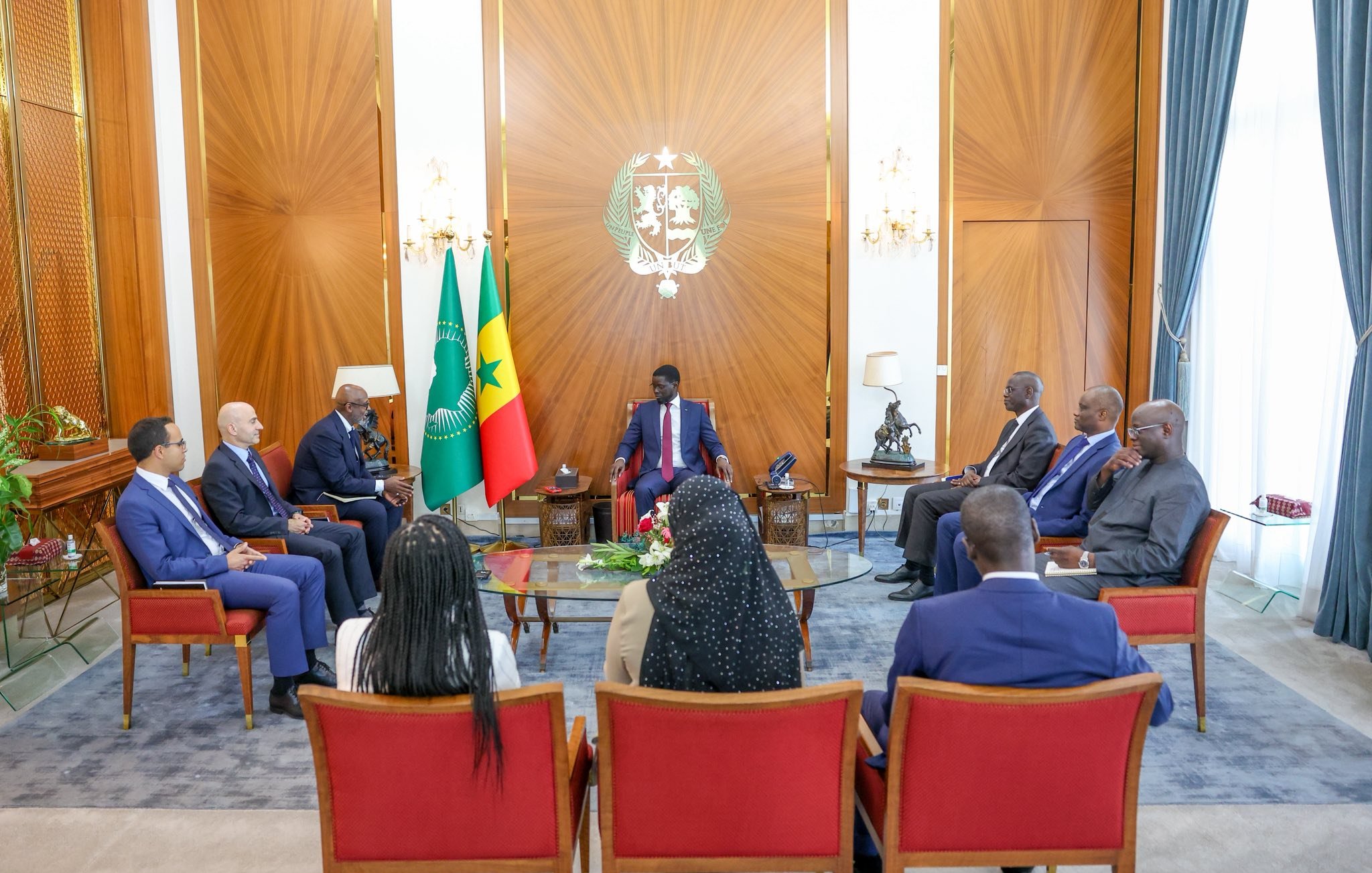 L'image montre une réunion dans une salle élégante. Au centre, un homme assis porte un costume et semble être à la tête de la discussion. Autour de lui, plusieurs personnes assisent dans des fauteuils confortables. À l'arrière-plan, on peut voir les drapeaux du Cameroun et du Sénégal. La pièce est lumineuse, avec des rideaux drapés et un décor raffiné, donnant une atmosphère formelle et professionnelle à cette rencontre. Les participants semblent engagés dans une conversation sérieuse.