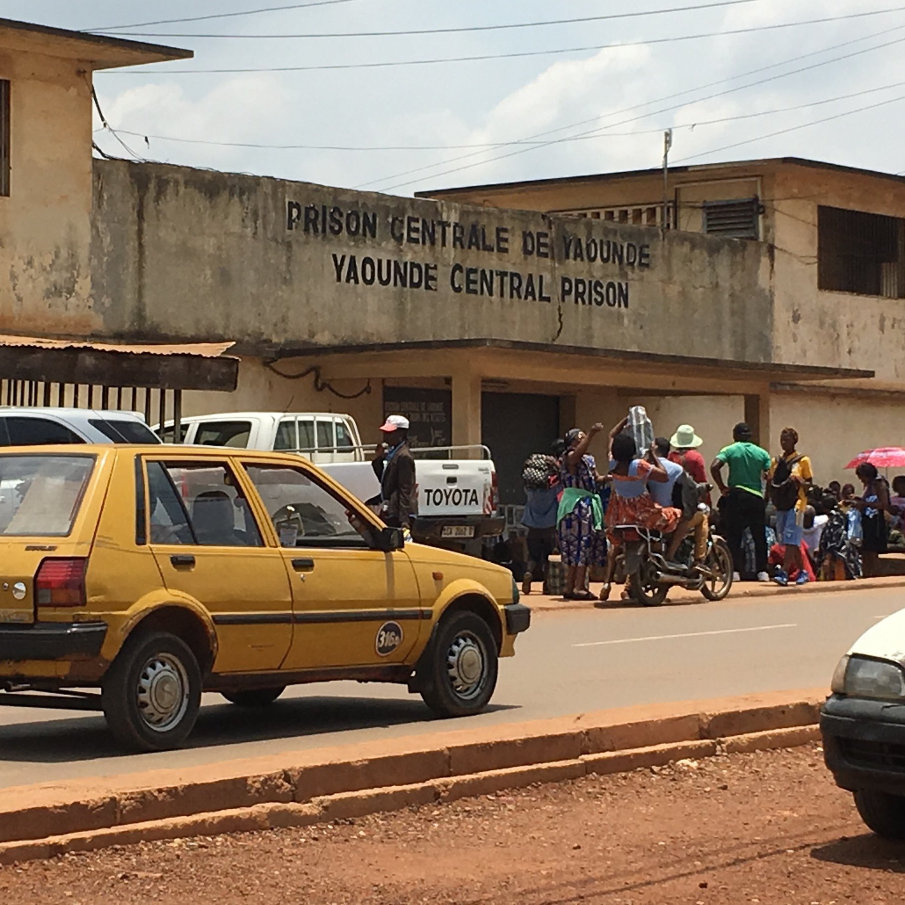 L'image montre l'extérieur de la prison centrale de Yaoundé, au Cameroun. On peut voir un bâtiment sobre avec des murs en béton, portant l'inscription "PRISON CENTRALE DE YAOUNDÉ" en lettres visibles sur la façade. En face de la prison, une route est occupée par une voiture jaune, typique des véhicules urbains. À côté, plusieurs personnes sont rassemblées, certaines discutant, d'autres attendant. Le décor autour est urbain, typique des villes africaines, avec une atmosphère animée. Le ciel est partiellement nuageux, ajoutant une nuance à la lumière de la scène.